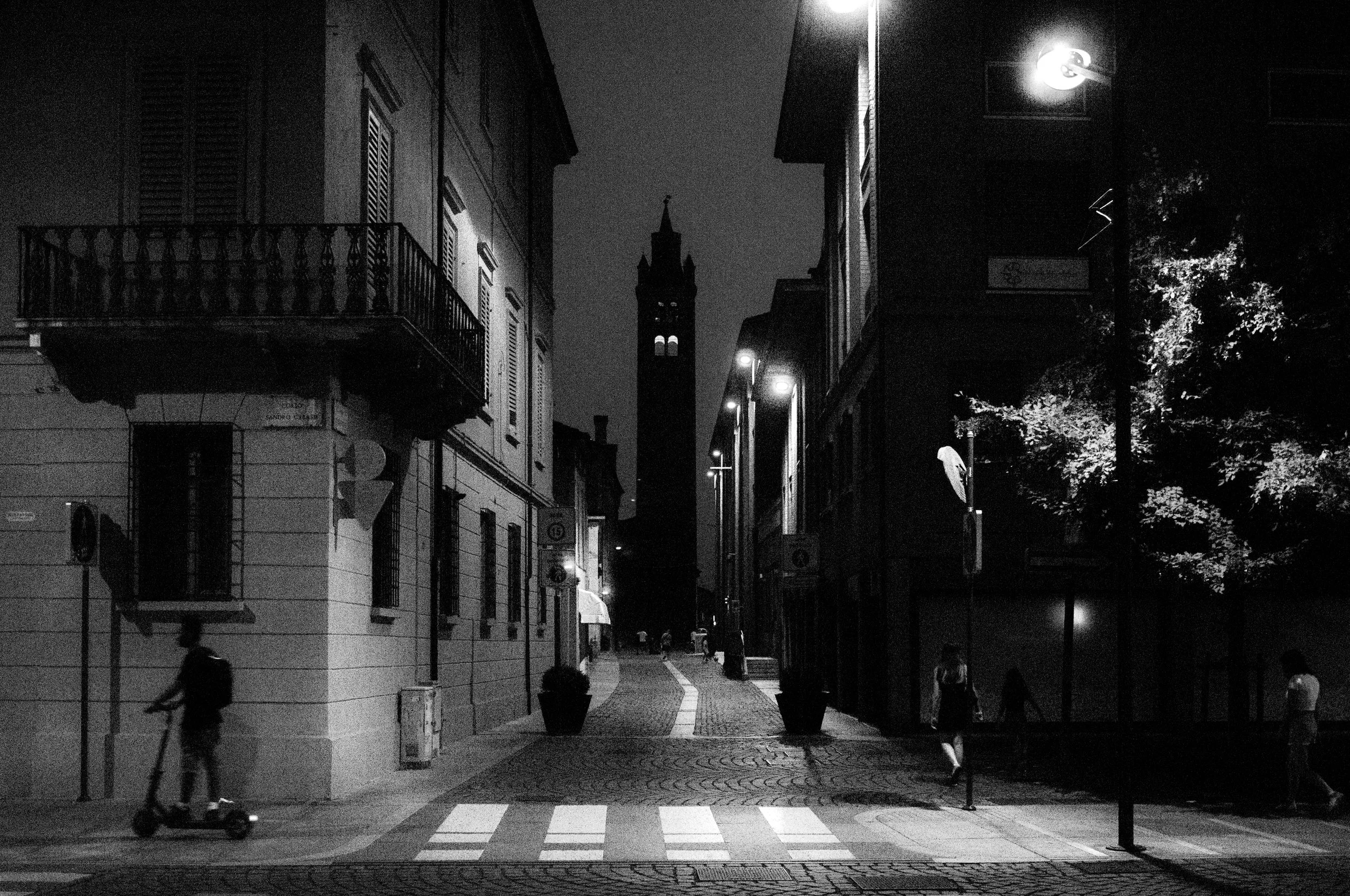 Una strada di città di notte con edifici storici, luci stradali e una torre con orologio sullo sfondo, alcune persone e un monopattino elettrico in movimento.