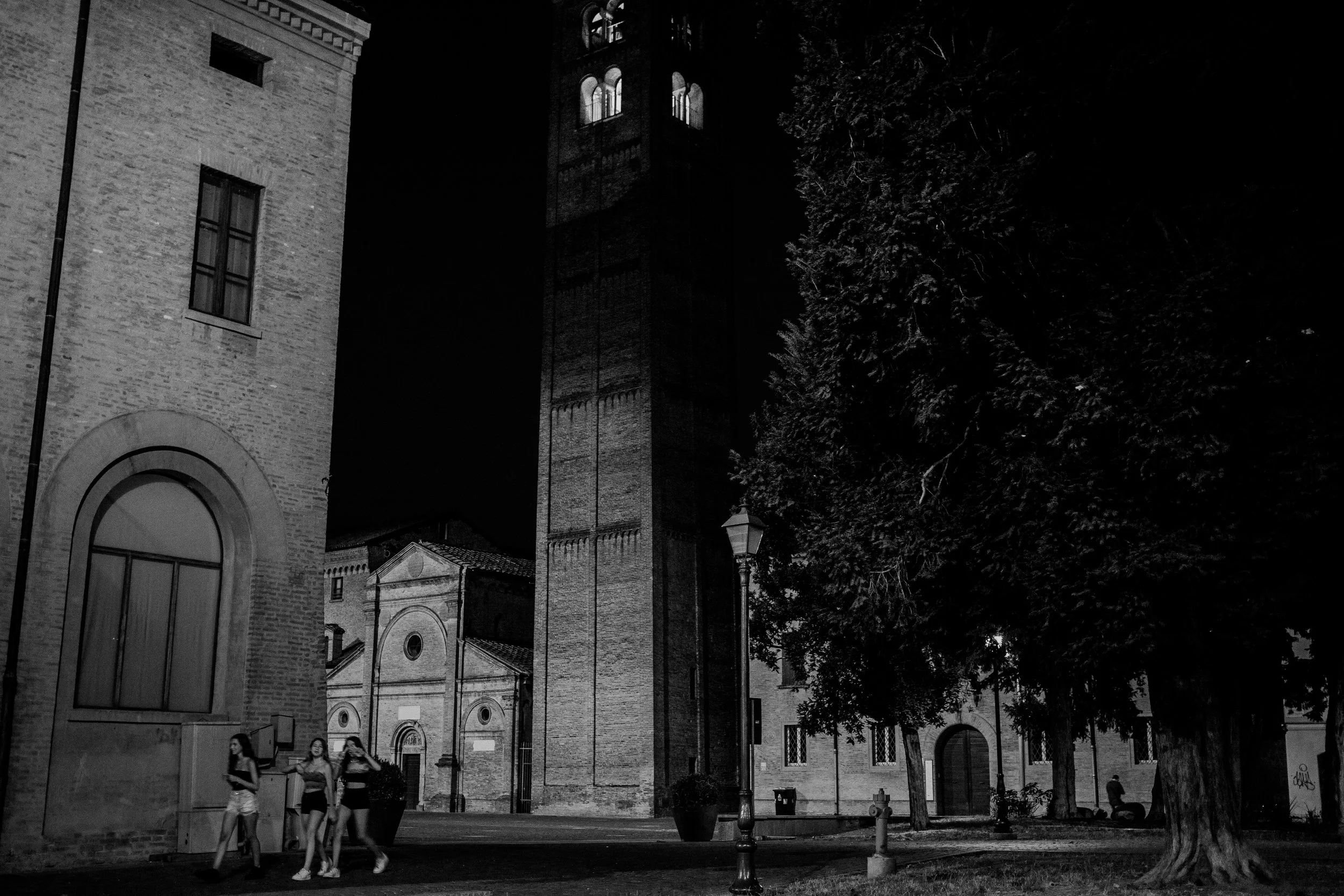 Scena notturna in un centro storico, con un campanile in mattoni, alberi e alcune persone camminando sulla strada.
