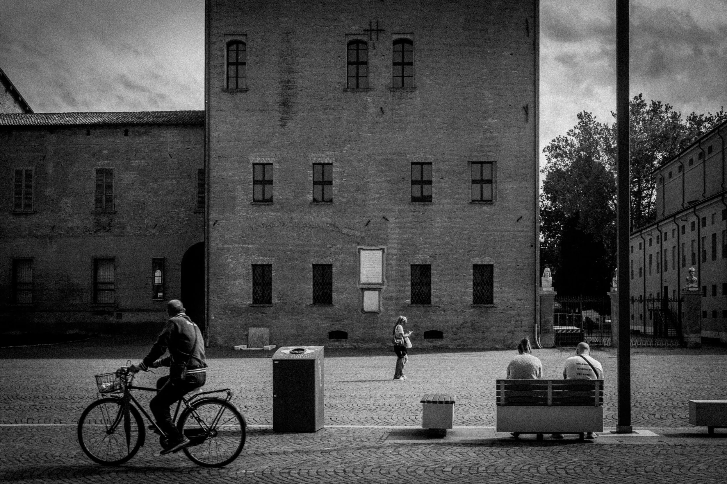 Persone che si rilassano e una in bicicletta in una piazza di fronte a un grande edificio di mattoni, in bianco e nero