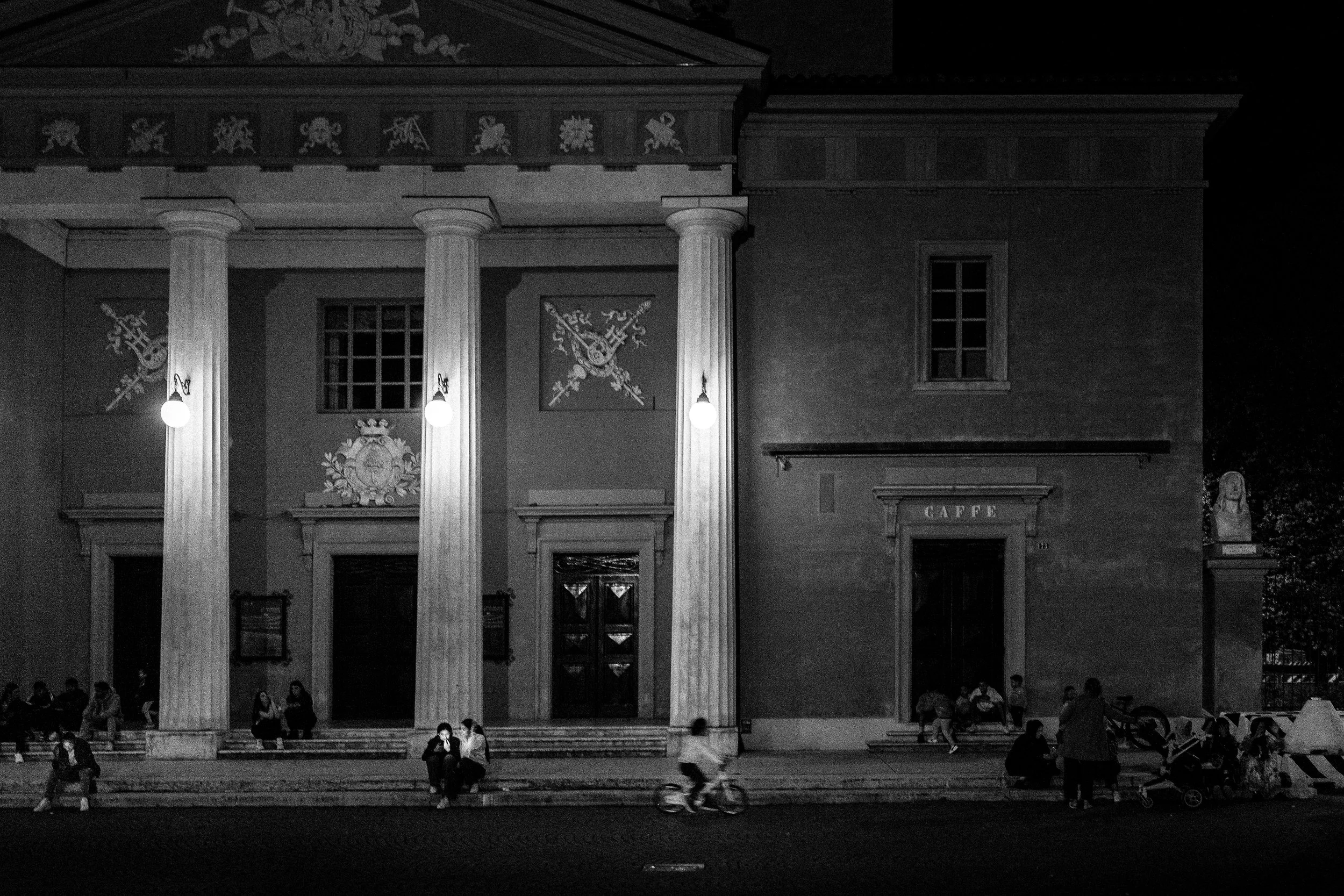 Immagine di un edificio storico con colonne e decorazioni, illuminato di notte, con persone sedute e alcune in bicicletta davanti. Signo 'Caffè' sulla porta.