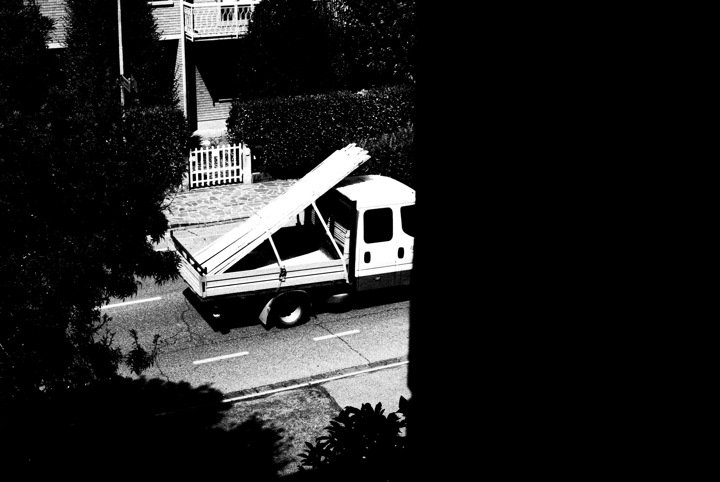 Un camioncino bianco con strada, cespugli e cancelli di recinzione visibili, parte di un edificio in alto. La maggior parte dell'immagine è nero, con una sezione centrale in bianco e nero della strada e del camion.