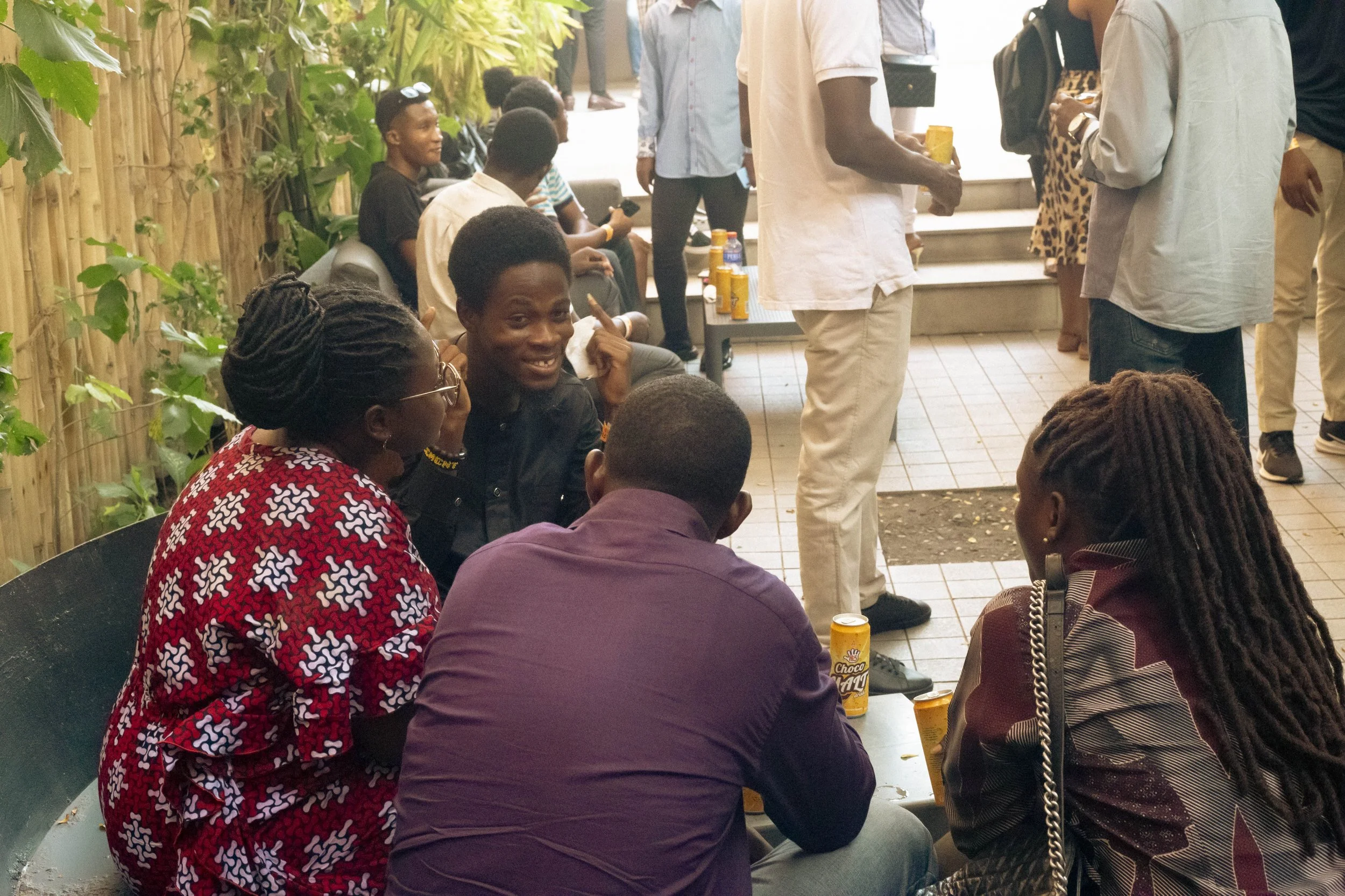 Group of African American people socializing outdoors, sitting and talking at a table with snacks and drinks, while others stand and chat in the background.