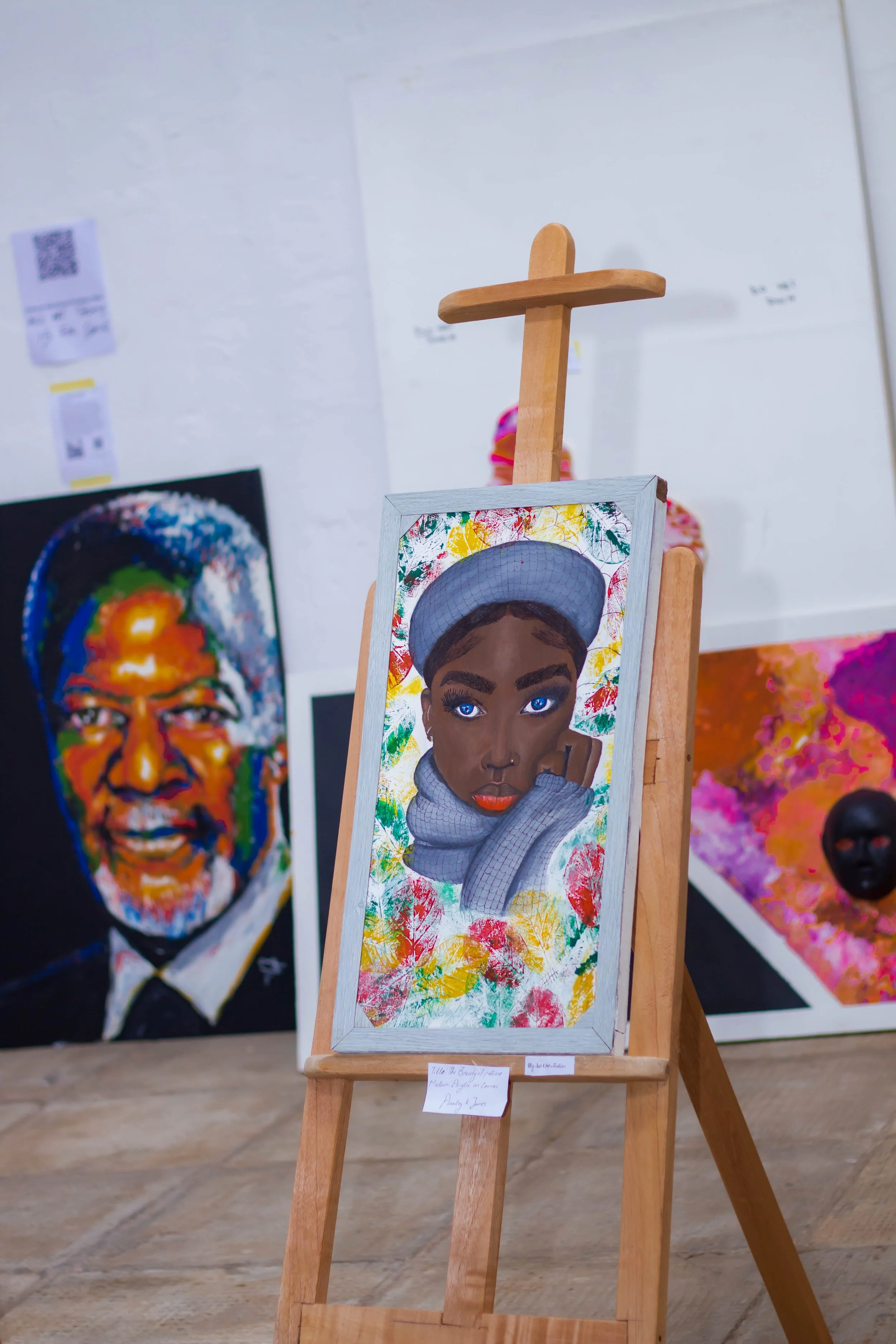 A colorful portrait of a young woman with dark skin, wearing a gray hoodie and a cap, displayed on an easel at an art exhibition. In the background, there are other vibrant artworks, including a portrait of a smiling man and a colorful abstract piece