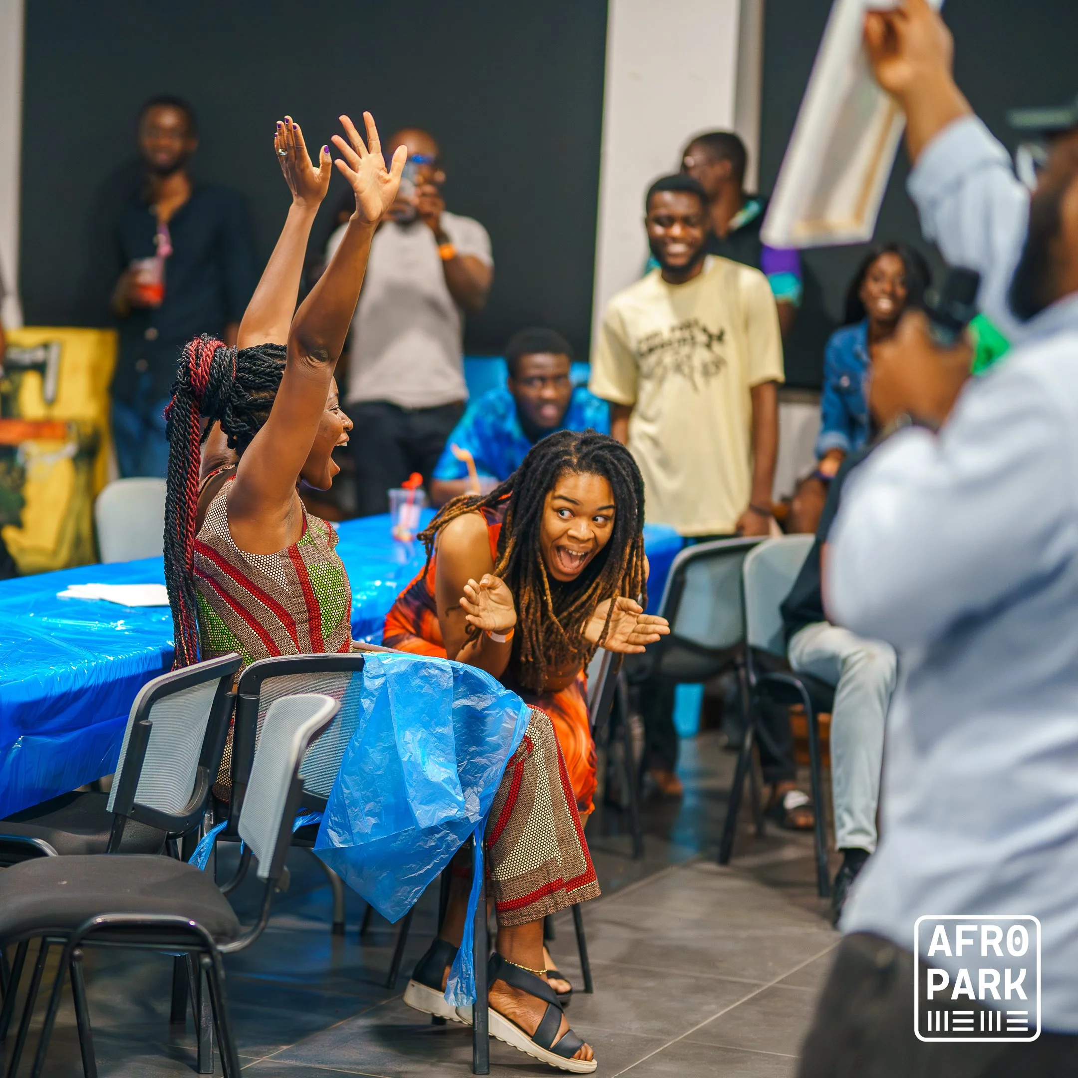 A lively group of people at a celebration, with two women at the center showing excitement and joy. One woman has her hands raised, and the other is clapping and smiling. Others in the background are also smiling and enjoying the moment, with some st