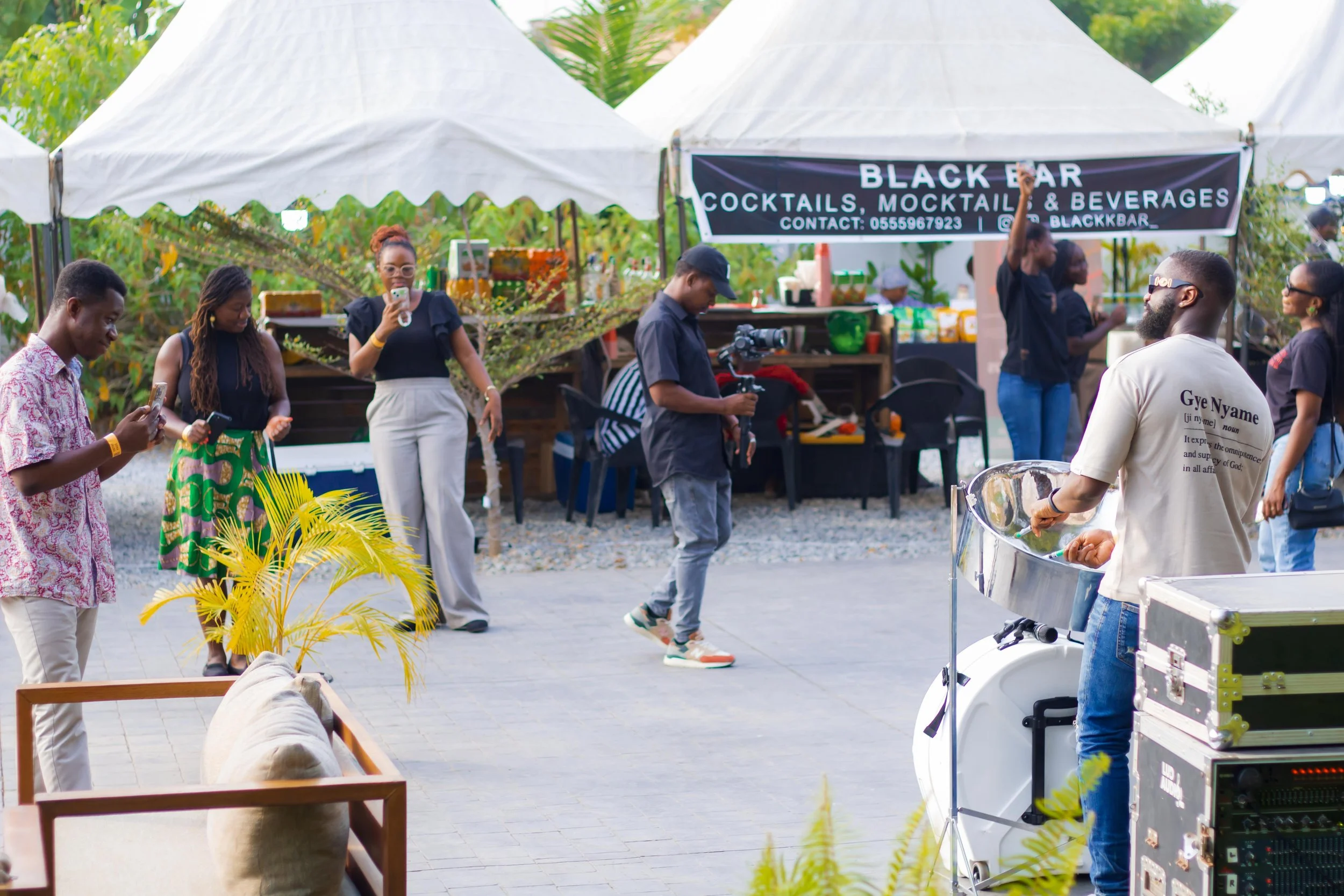 People enjoying a daytime outdoor event near a food stall with a banner that reads 'Black Bar Cocktails, Mocktails & Beverages.' Some individuals are using their phones, and others are observing or taking photos. There is a musician or performer play