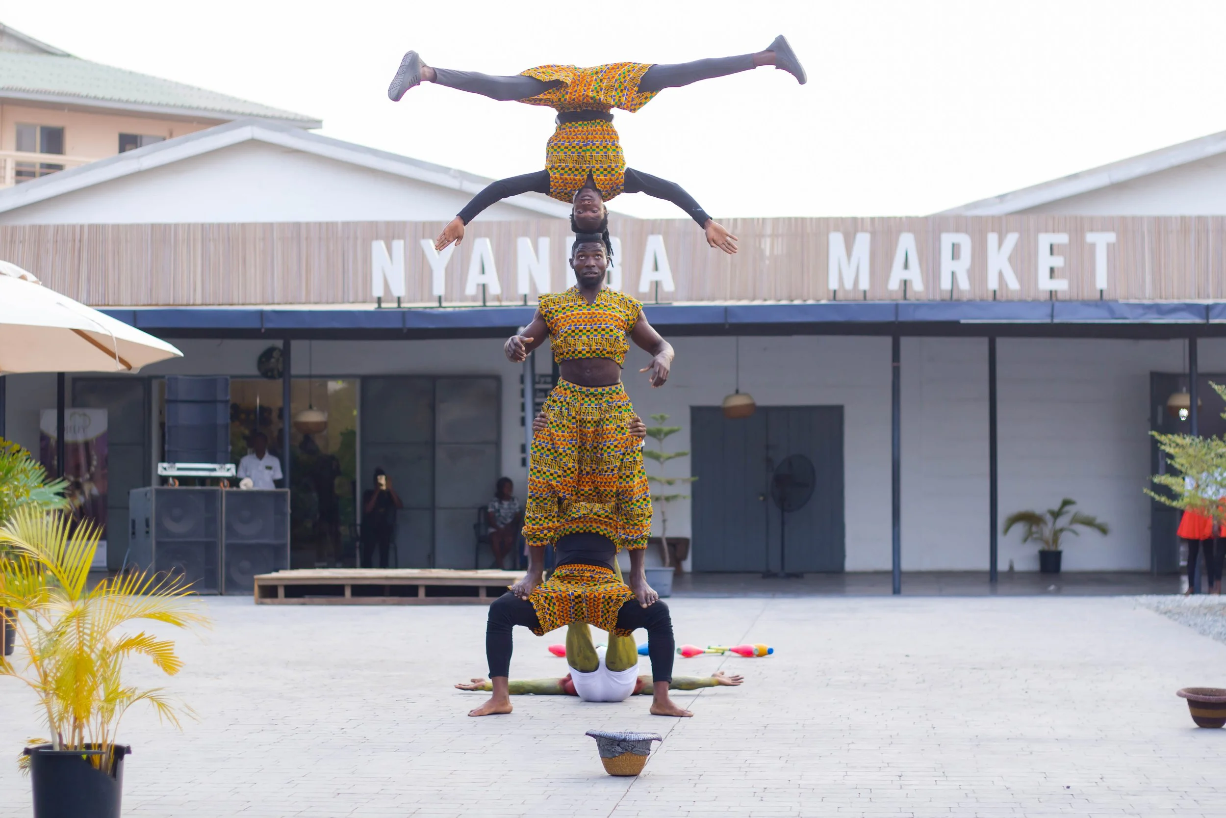 Four acrobats performing a human pyramid in front of a market, with the top acrobat doing a handstand, outside Nyanza Market building.