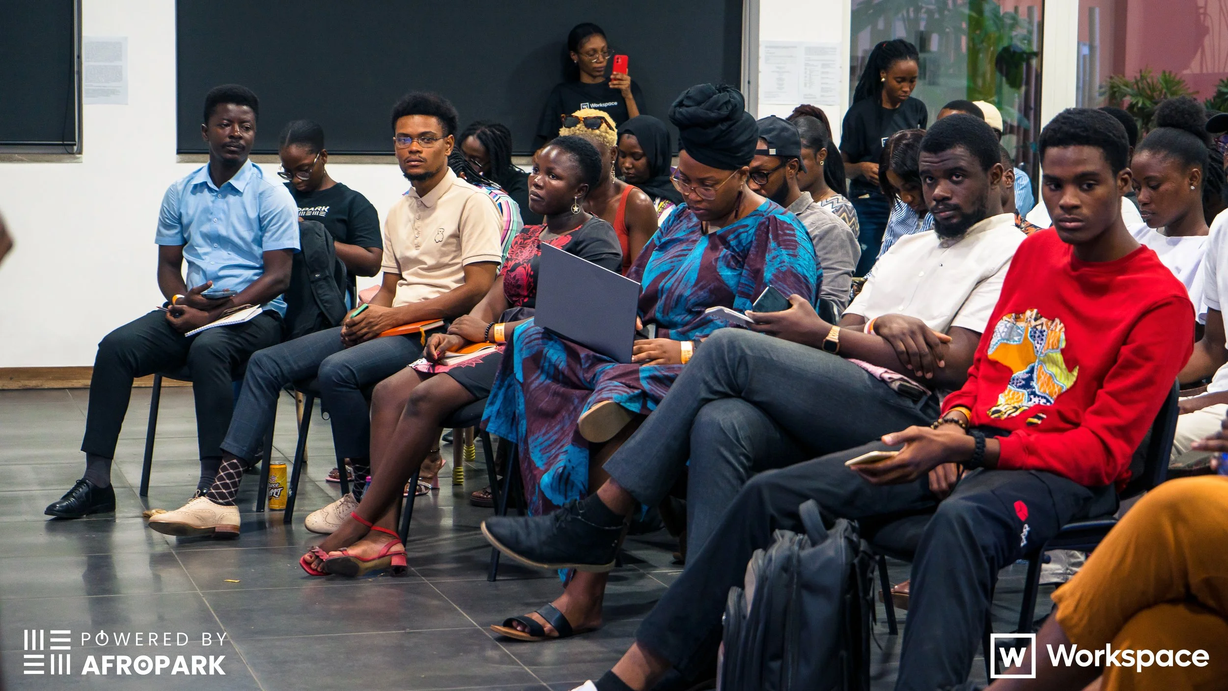 A group of diverse people seated in a conference or seminar room, some using laptops and phones, attentively listening to a presentation.