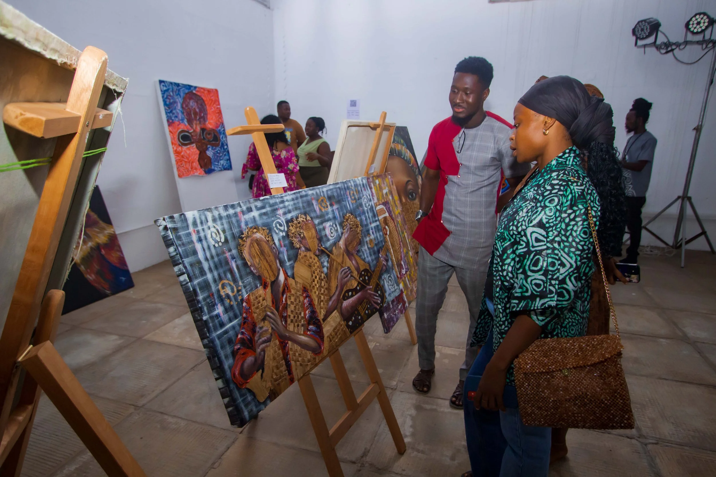 People viewing colorful artwork at an art gallery, with paintings on display and spotlights on the ceiling.