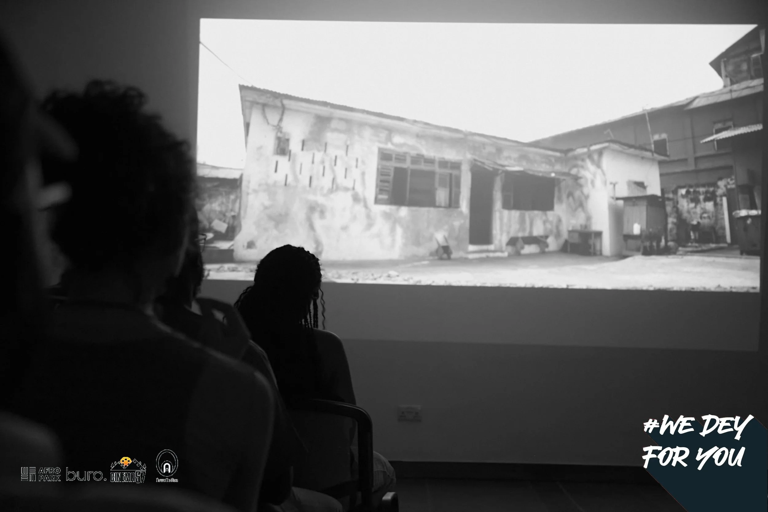 Silhouettes of two people watching a black and white video projected on a wall, showing an old building with a worn exterior and damaged walls. The scene includes chairs and a dark room setting. Text in the lower right corner reads '#WE DEY FOR YOU'.