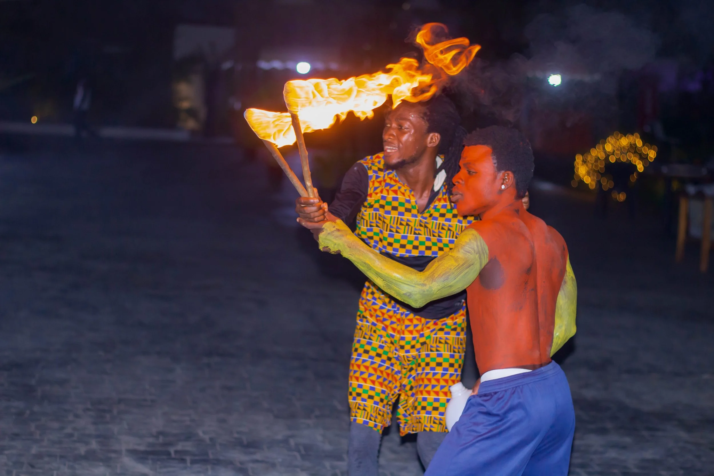 Two men performing a fire dance outdoors at night, with one man holding fire sticks and the other with painted body, wearing colorful clothing.