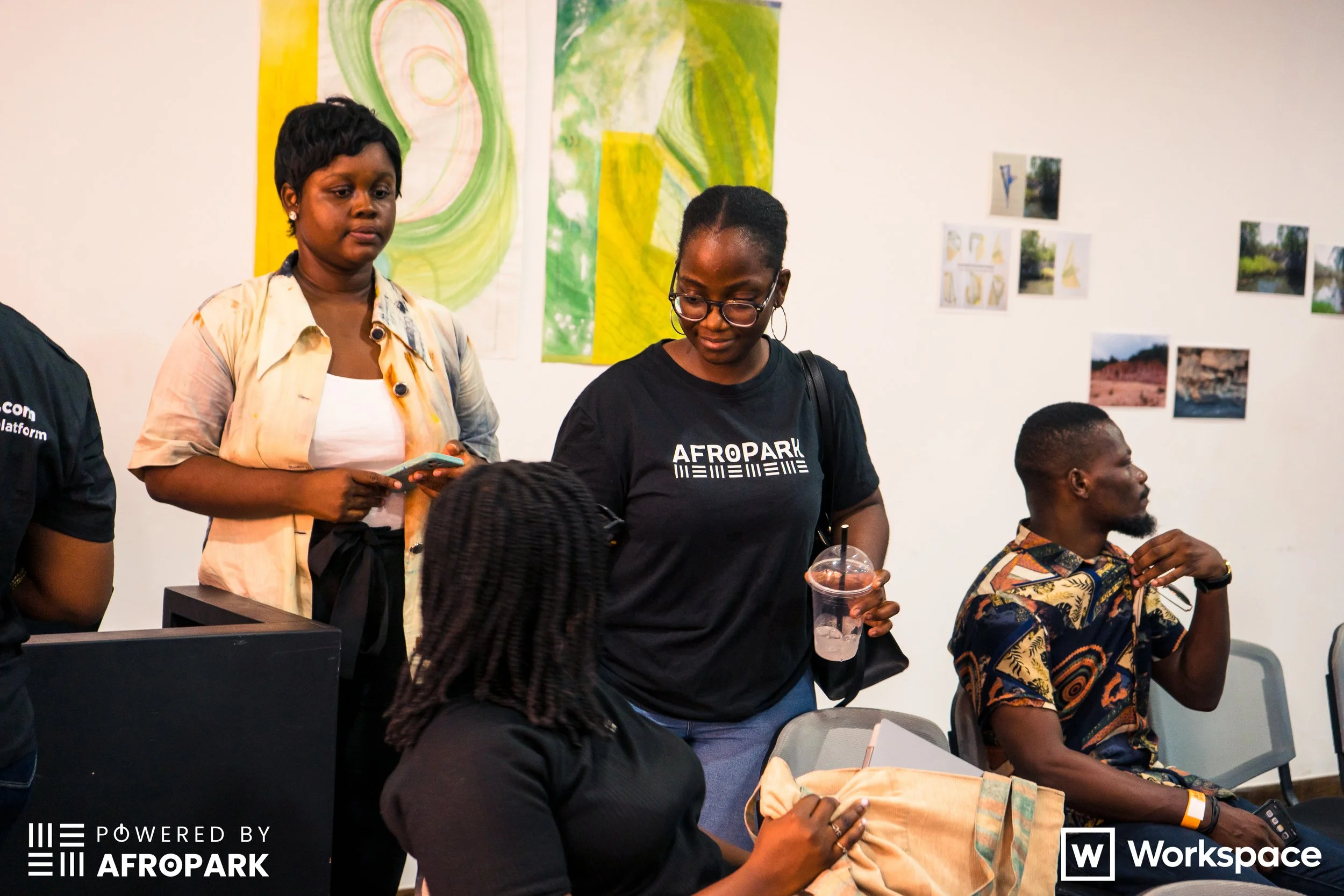 Group of four people in a room decorated with abstract and nature-themed artwork. One woman with glasses and a black t-shirt labeled "AFROPARK" stands holding a drink, another woman with short hair and a beige shirt stands looking at her phone, and a