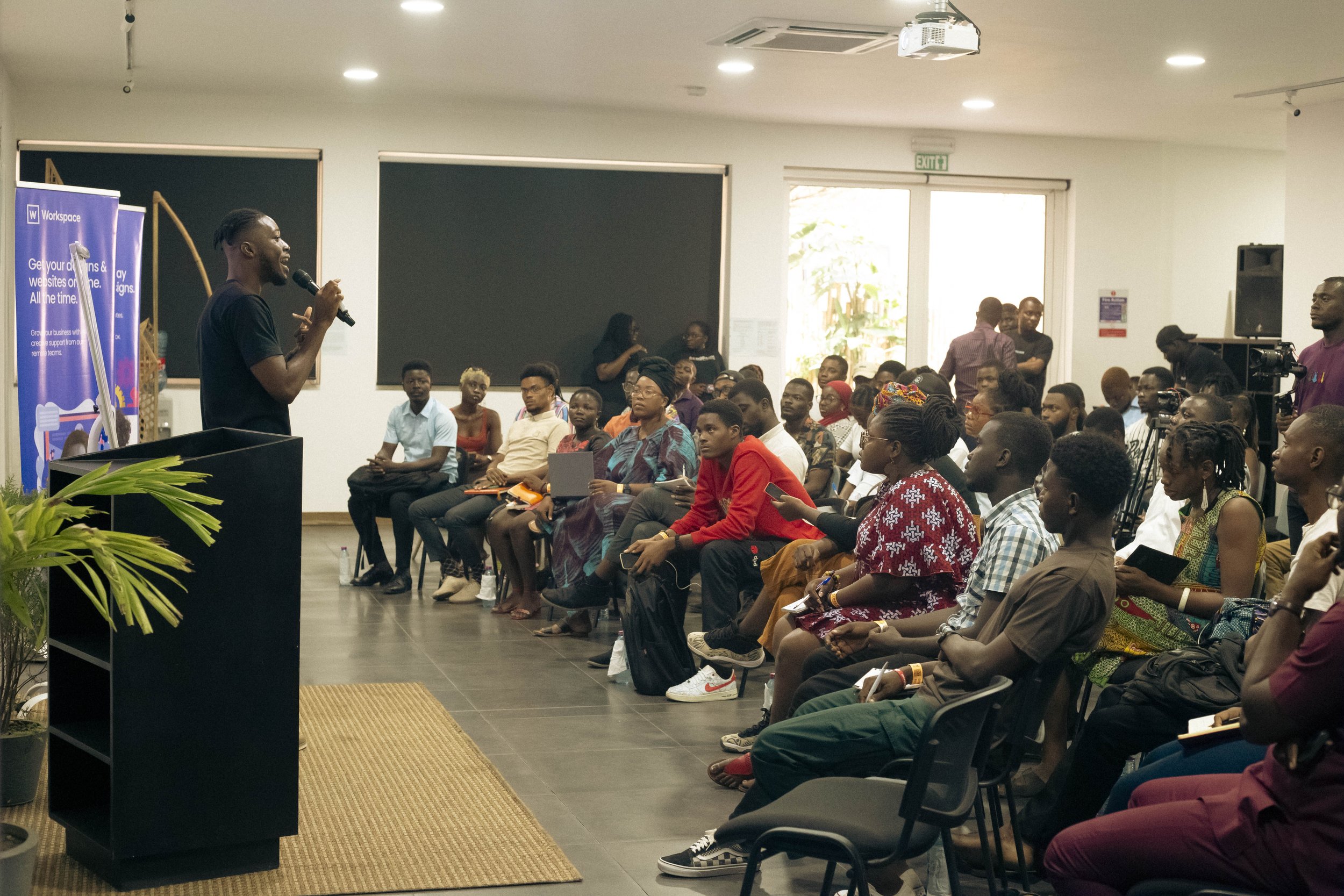 A man is speaking into a microphone at the front of a room full of seated people, during a presentation or seminar. The audience appears attentive, with some taking notes or using devices. There are blackboards and a banner on the side.