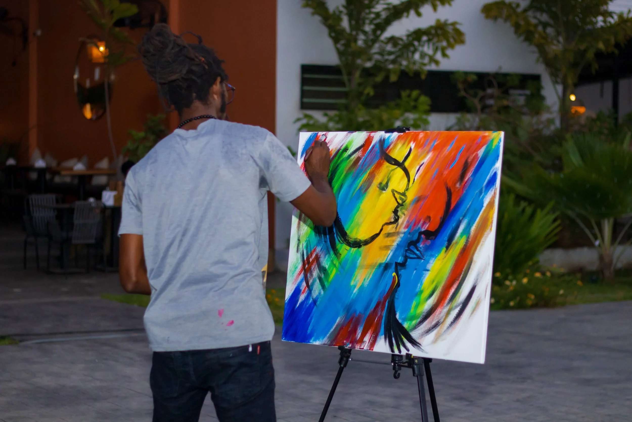 A person with dreadlocks and glasses painting an abstract colorful artwork on a canvas outdoors during dusk.