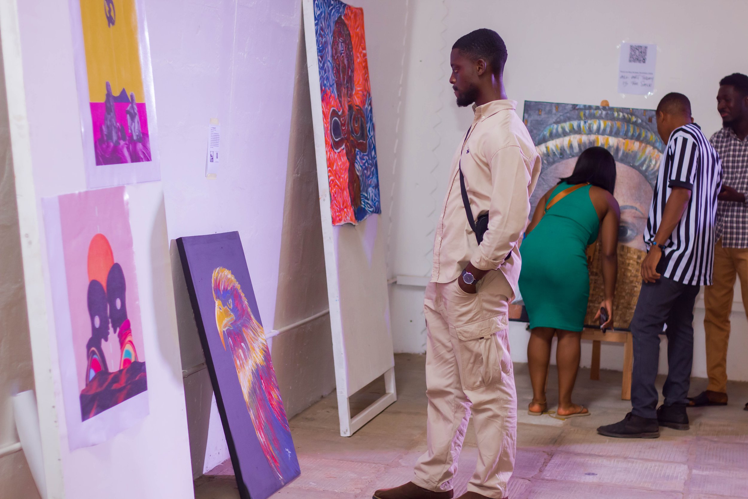 People observing artwork in an art gallery, including paintings of abstract and vivid designs, with one person in a beige jumpsuit and others in casual attire.