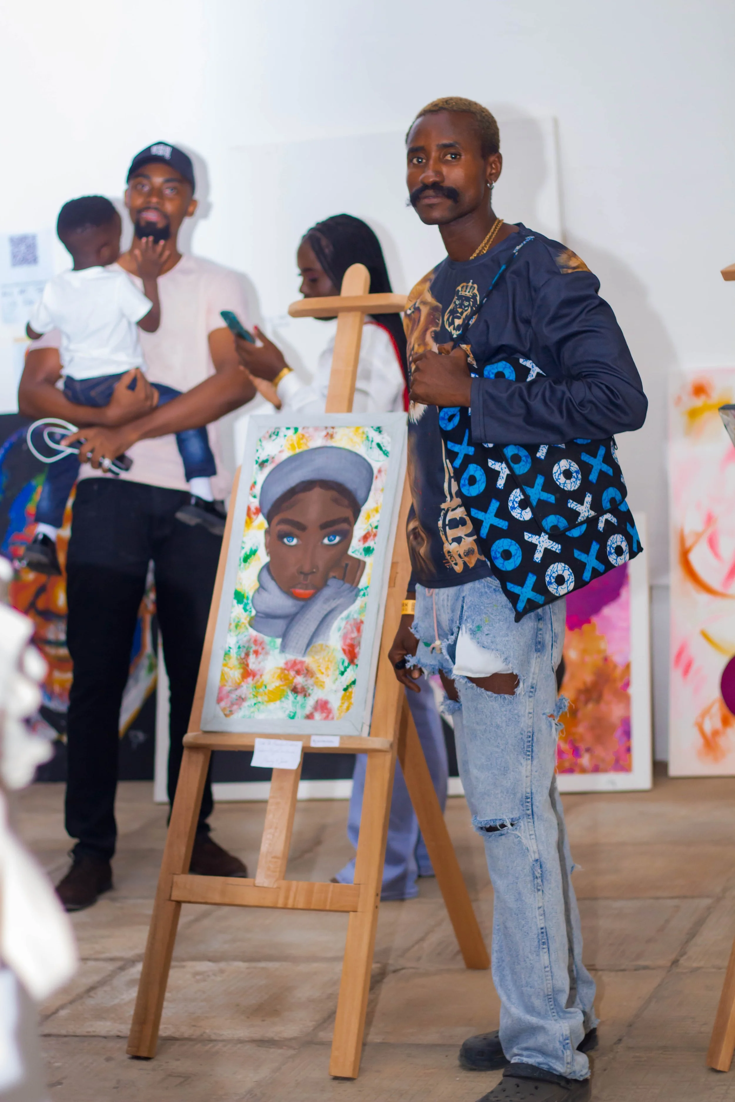 Man standing next to an art piece of a woman with blue eyes, painted on a canvas set on a wooden easel at an art gallery. Other people are visible in the background.