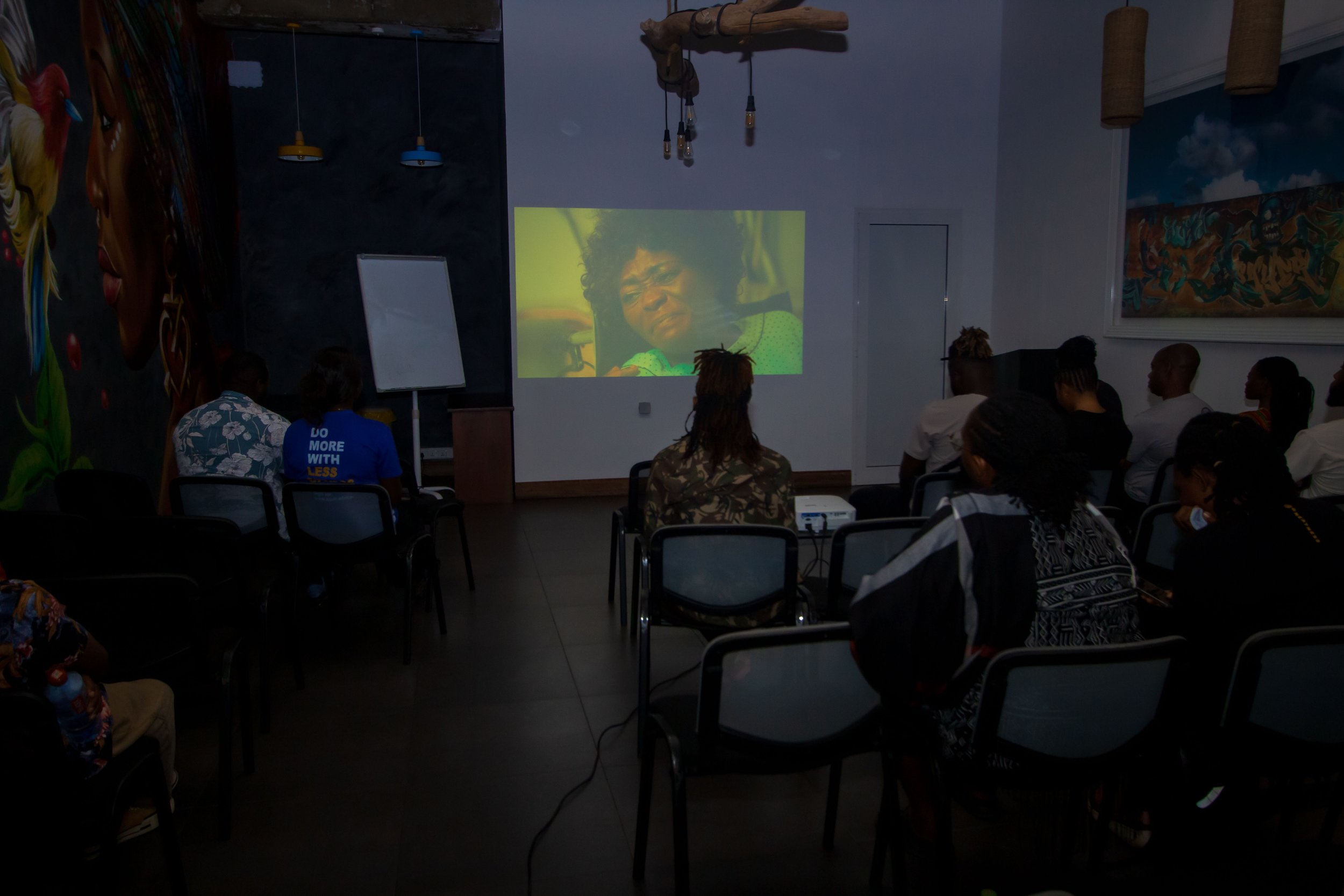 People sitting in a dimly lit room watching a movie or presentation projected on a wall. The room has artwork on the walls and chairs arranged facing the screen.