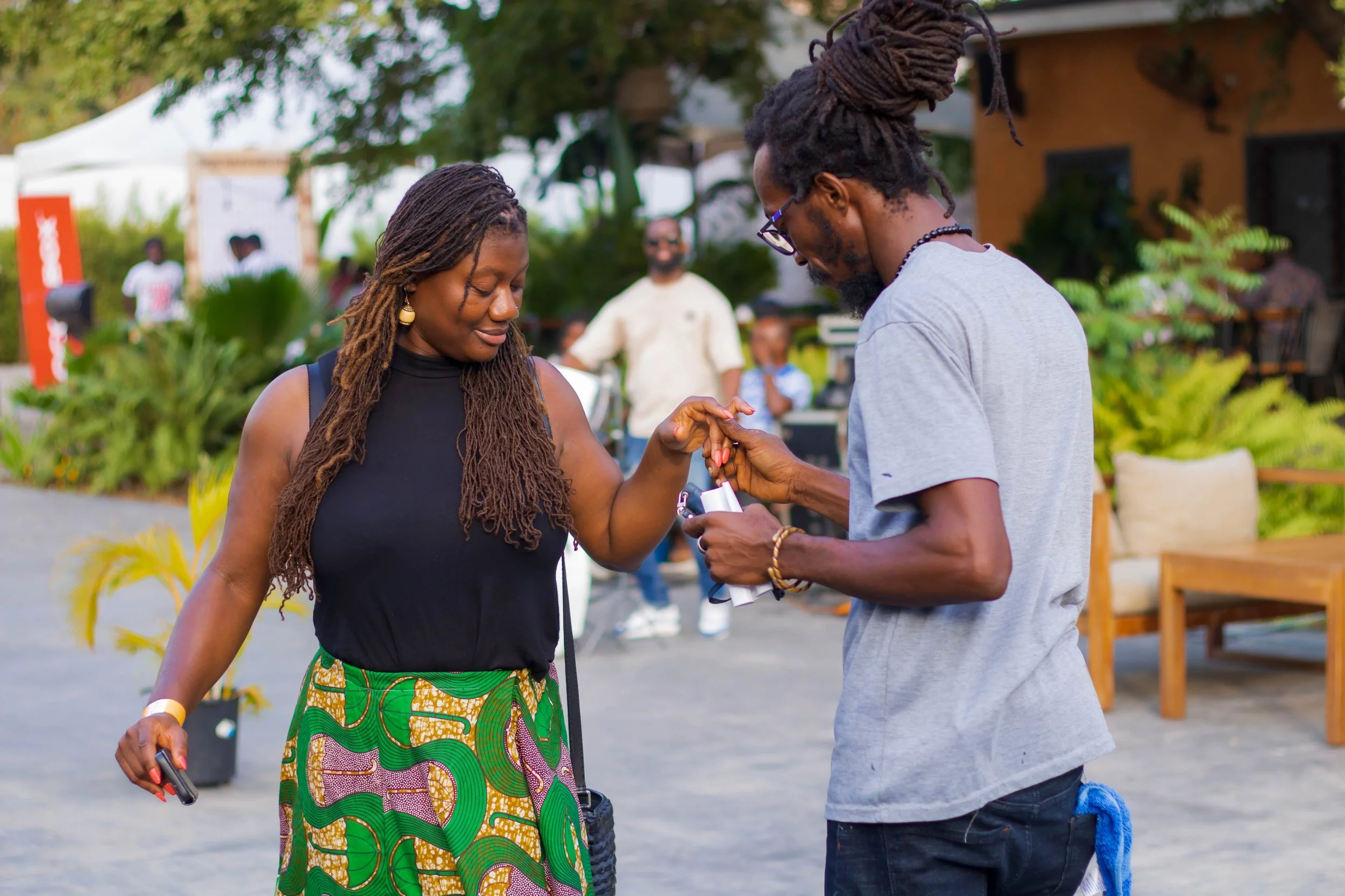 A woman in a black sleeveless top and colorful skirt smiles as a man in a gray t-shirt and glasses puts a ring on her finger during an outdoor event.