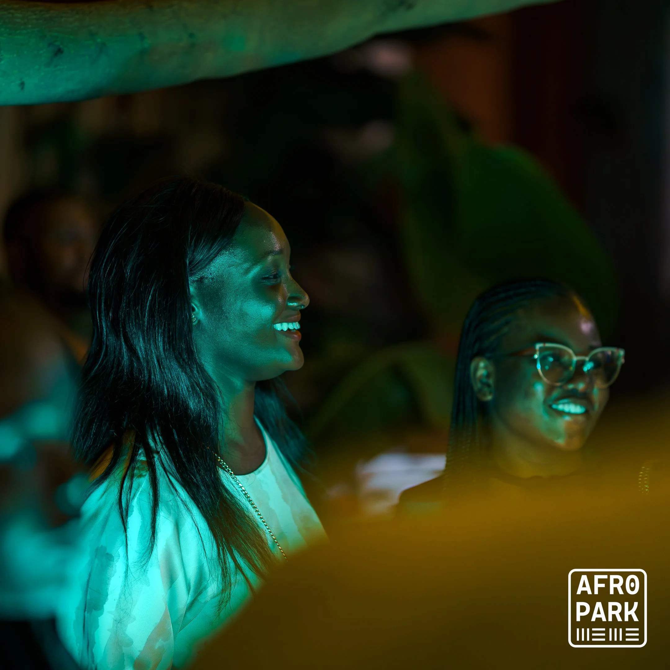 Smiling woman with long dark hair and woman with glasses in a dimly lit setting, illuminated by a greenish-blue light, at Afro Park event.