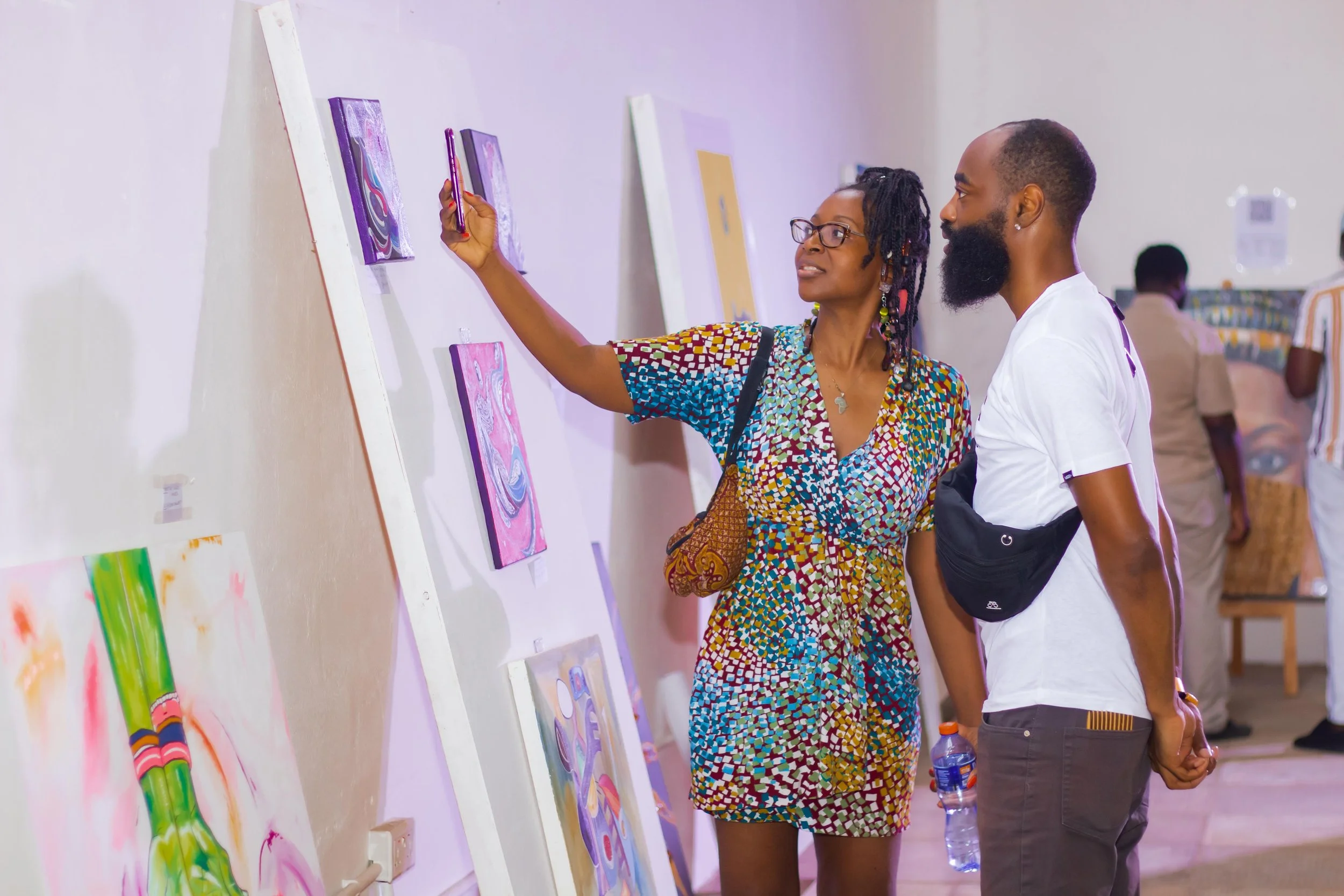 A woman and a man are viewing artwork on a gallery wall. The woman is pointing at a piece and holding a phone; she has glasses and is wearing a multicolored patterned dress. The man is holding a water bottle and has a camera bag around his waist.