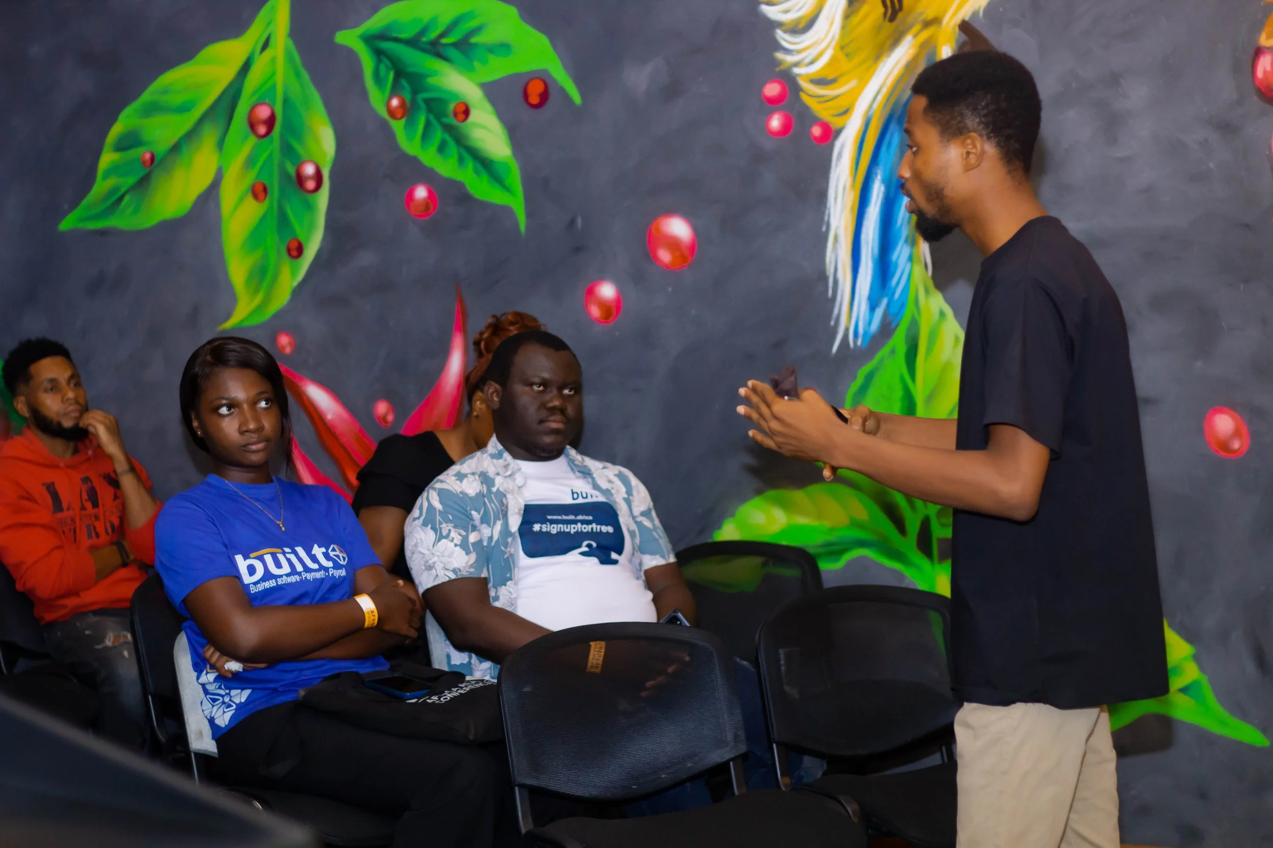 A young man giving a presentation to a small audience seated in a room with a colorful painted mural of coffee leaves and beans on the wall.