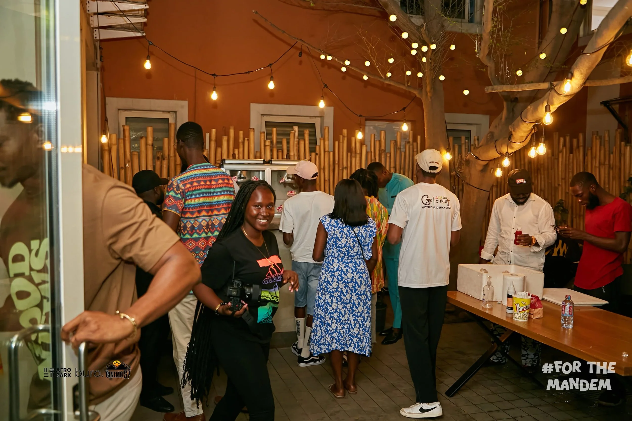 People gathered at an outdoor evening event with string lights hanging from trees. Some are preparing food at a counter, while others are socializing. A woman in the foreground is smiling at the camera, holding a camera. The setting has a relaxed atmosphere and is enclosed by a bamboo fence. The text '#FOR THE MANDEM' is visible in the bottom right corner.