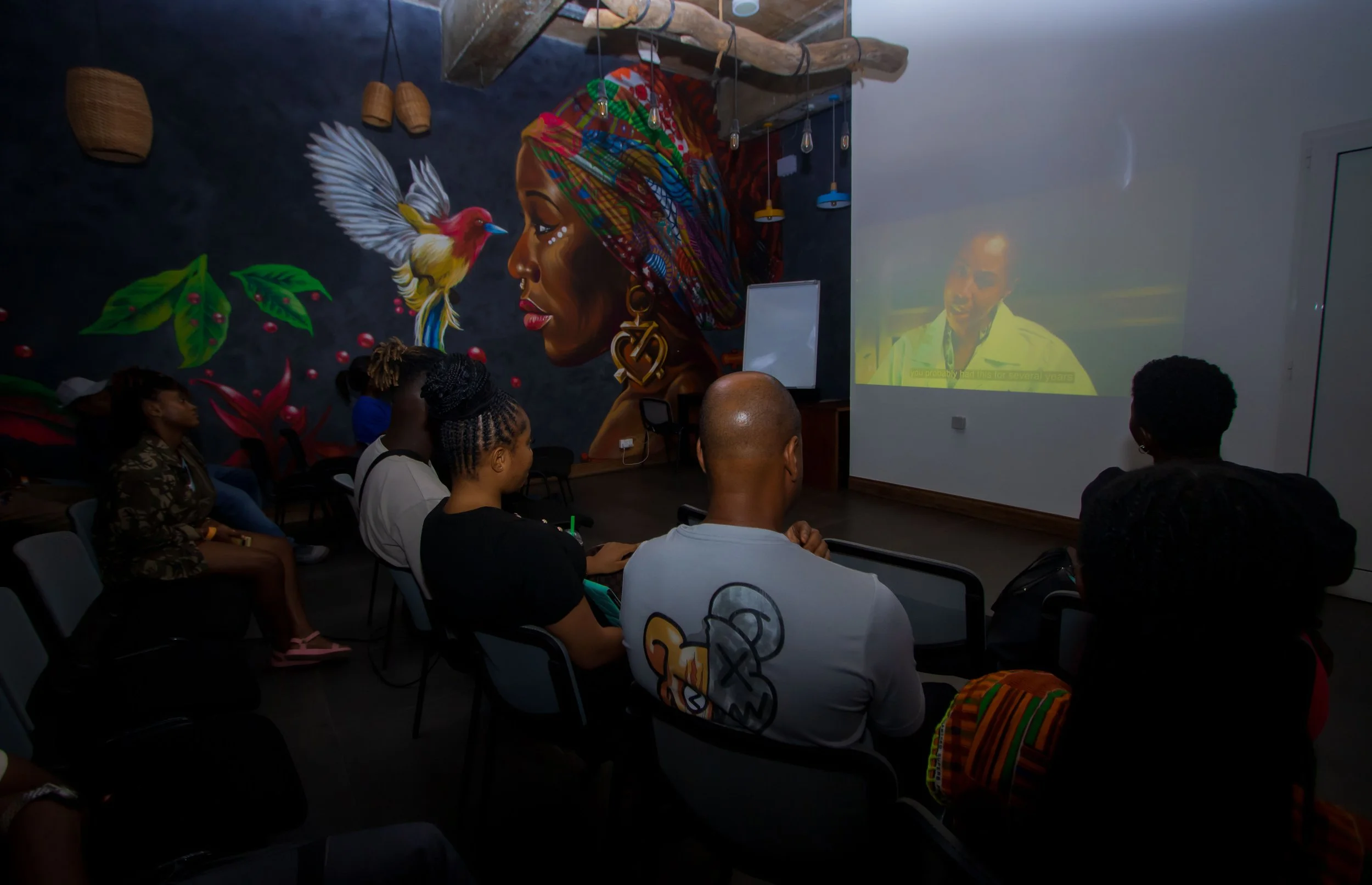 People watching a video in a dark room with a colorful wall mural depicting a woman's profile, a bird, and large leaves.