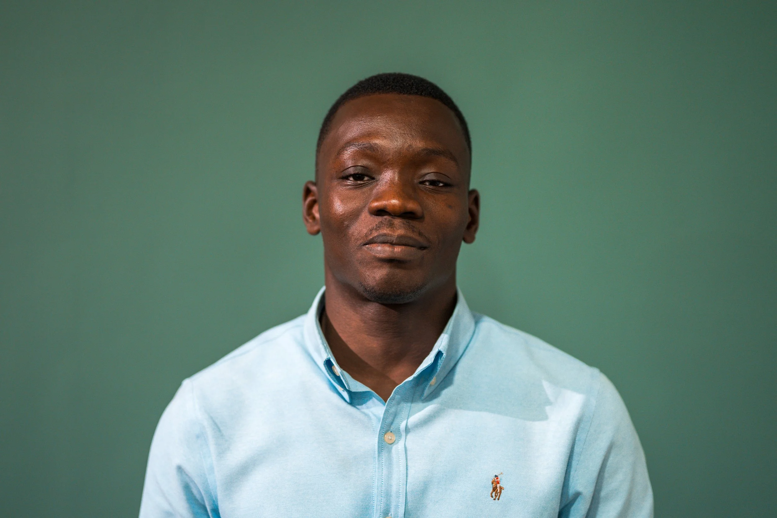 A young man with dark skin and short black hair is wearing a light blue collared shirt against a green background. He has a neutral expression.