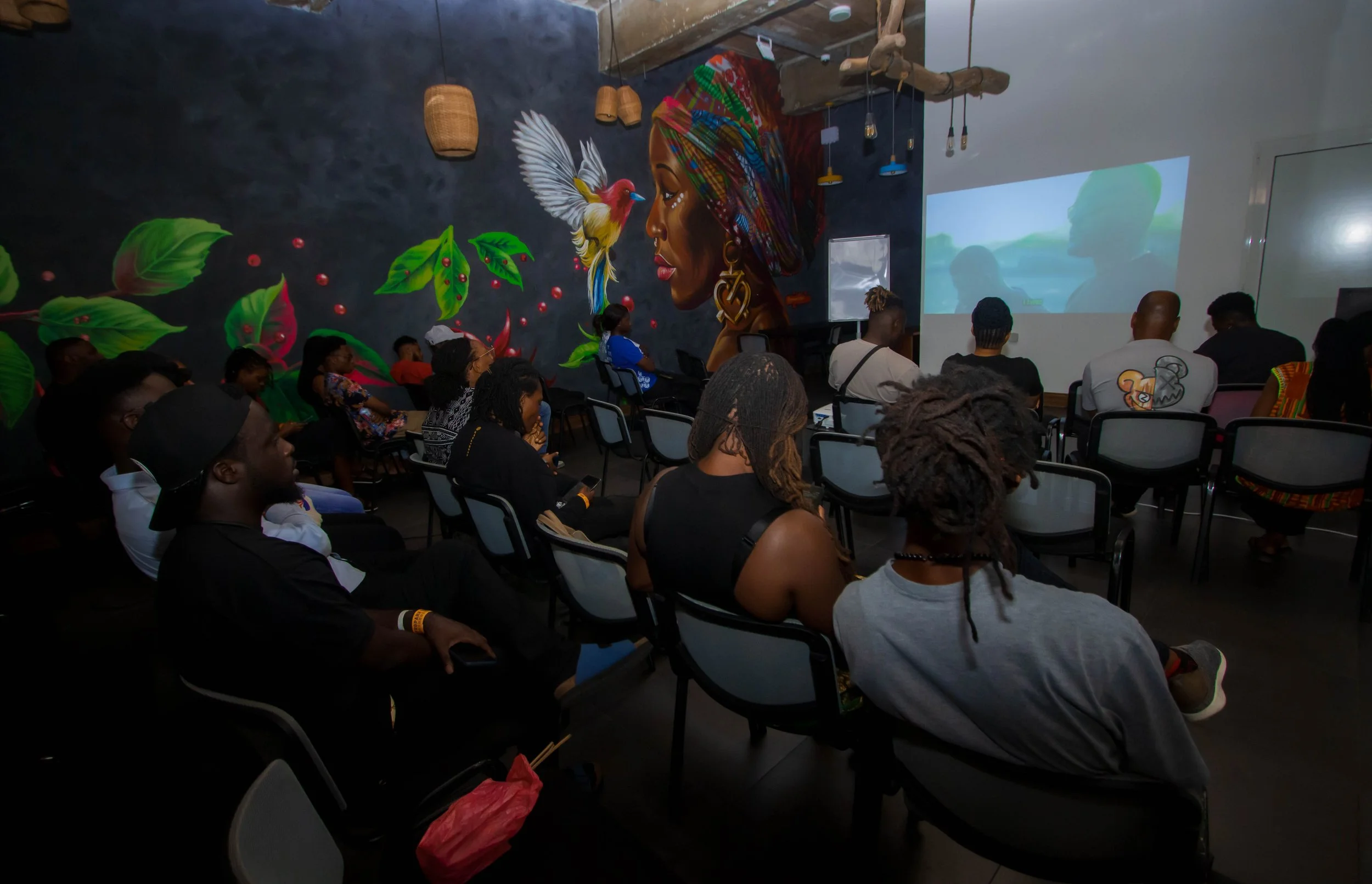 People seated in a dark room watching a movie projection on a white wall, with a colorful mural of a woman and a bird on the wall.