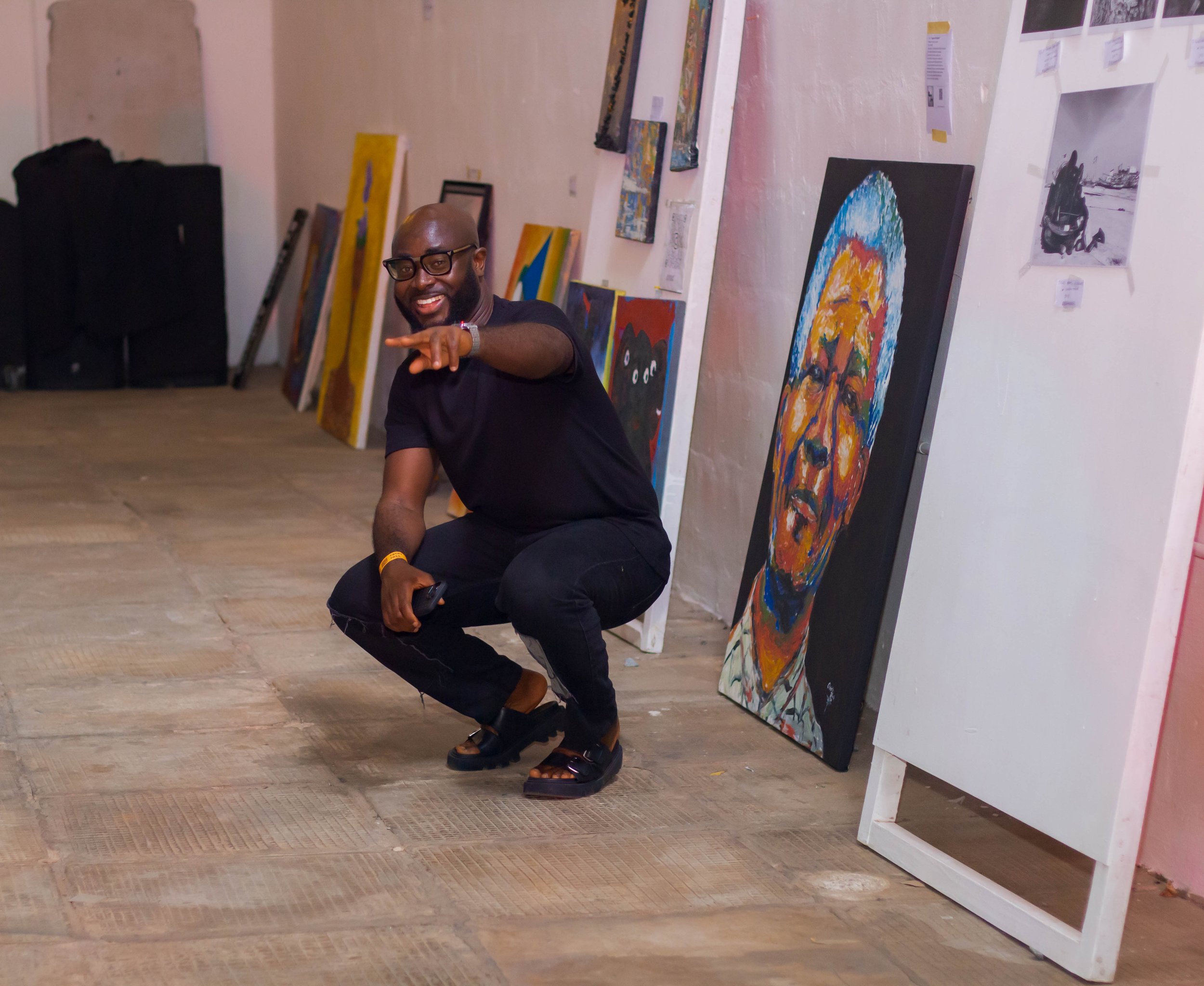 A man crouching and pointing at the camera in an art gallery with colorful paintings on the wall behind him.