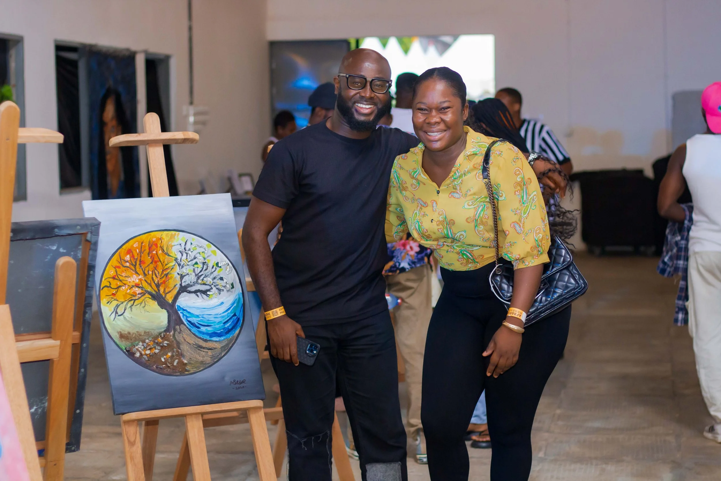 A man and woman smiling at an art exhibition, standing beside a circular painting of a tree with autumn leaves, a river, and a sky, displayed on an easel.