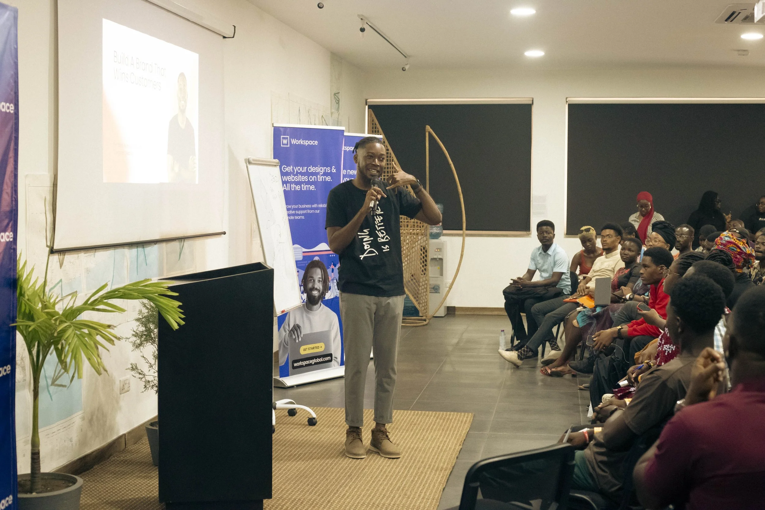 A man speaking into a microphone in front of an audience during a presentation. He is standing on a small rug, next to a laptop and a banner that reads 'Workspace'. The audience is seated and attentively listening, with some taking notes or photos. T