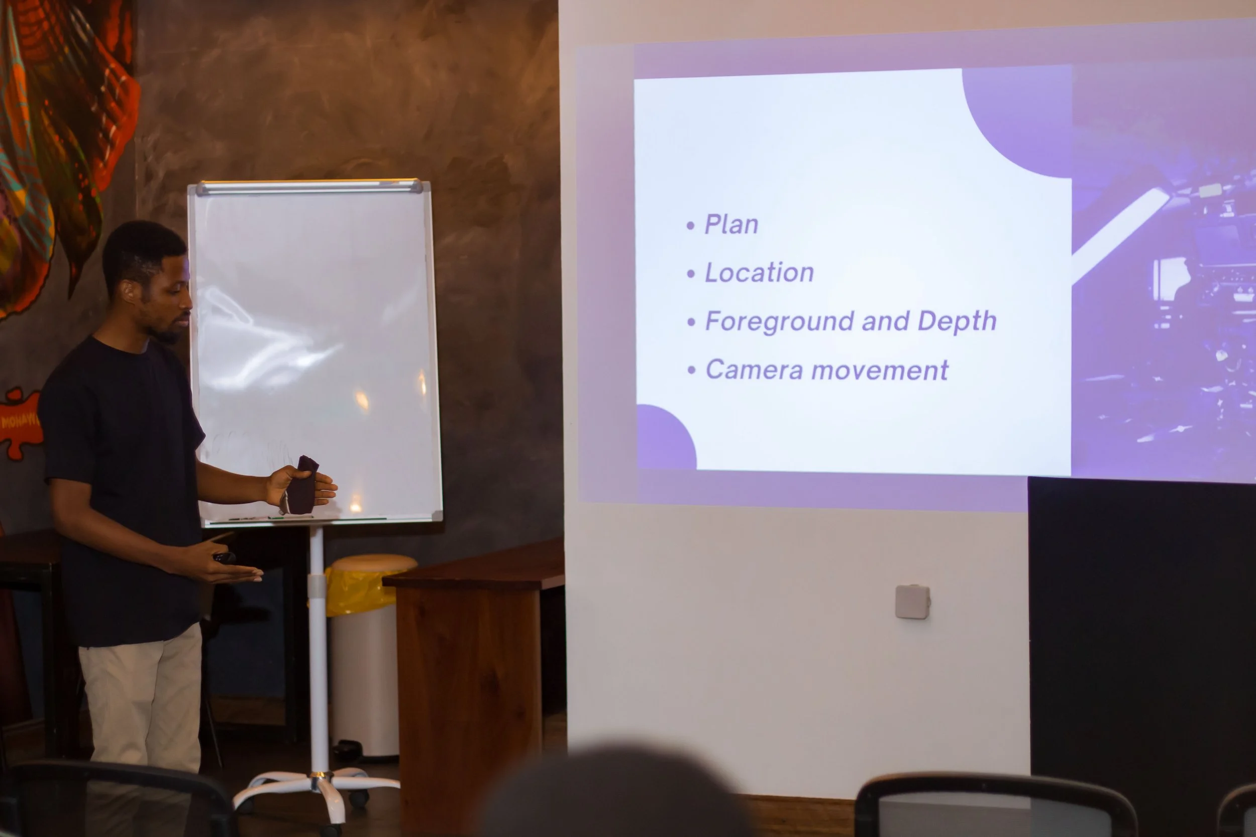 A man standing in a room delivering a presentation. There is a whiteboard and a projection screen displaying a slide with bullet points: 'Plan', 'Location', 'Foreground and Depth', 'Camera movement'.