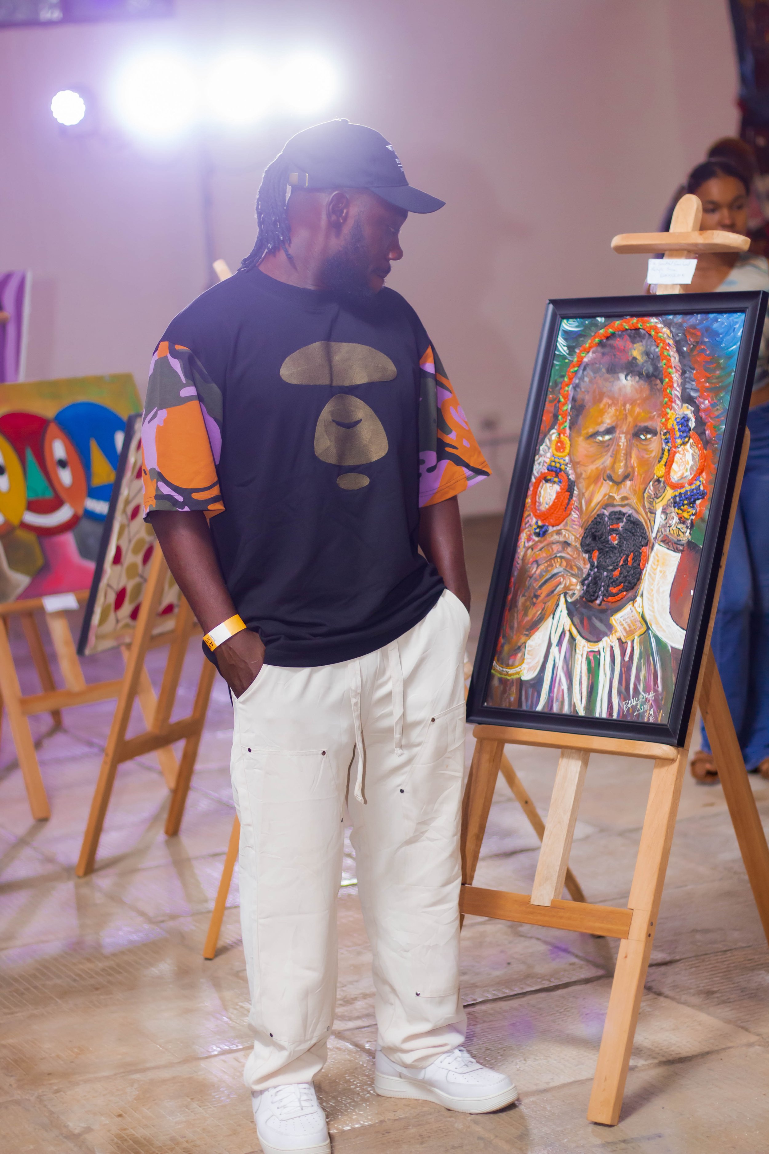 A man in a black T-shirt, white pants, and white sneakers is looking at a framed colorful portrait of a woman with beads and jewelry, displayed on an easel at an art gallery or exhibition.