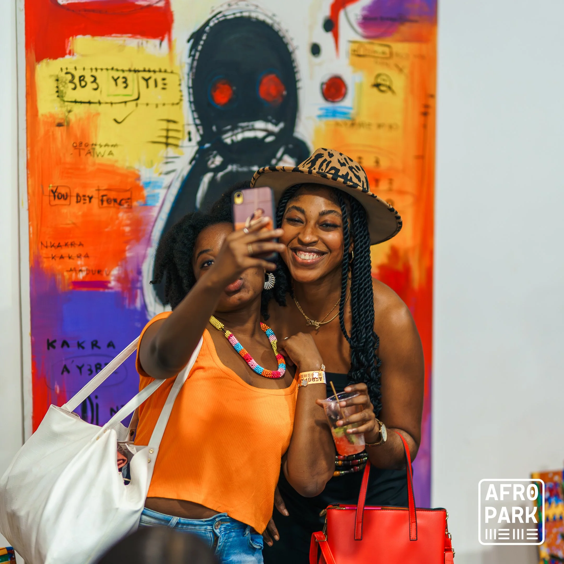 Two women taking a selfie at an art exhibit, with colorful abstract art in the background, one wearing an orange top, a beaded necklace, and a white bag, the other wearing a black top, a leopard print hat, and holding a drink and a red purse.