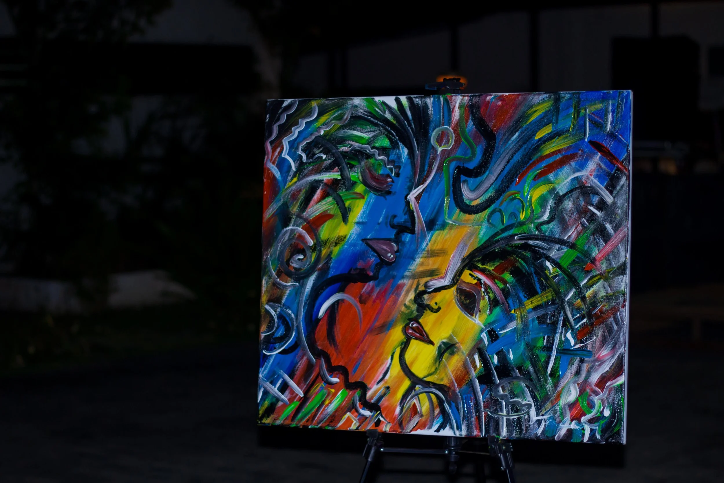 Abstract painting of multiple faces, with vibrant colors and swirling lines, displayed on an easel outdoors at night.