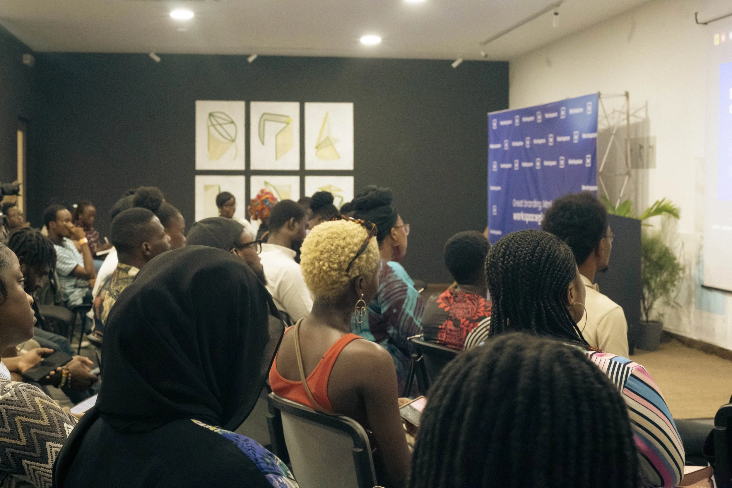 An audience of diverse people attending a presentation or lecture in a conference room, seated and looking toward a screen on the front wall.