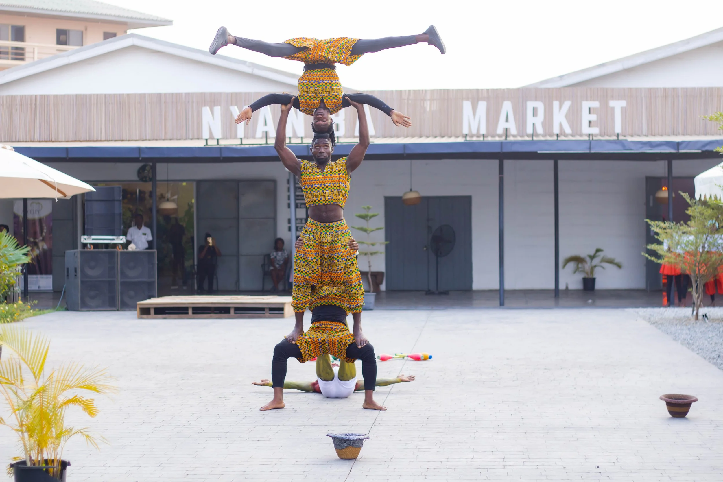 Four performers in colorful traditional African clothing are doing a human pyramid outside in front of a building with a sign that reads 'NANG MARKET.' One person is lying on the ground, supporting another in a handstand, while a third performer bala