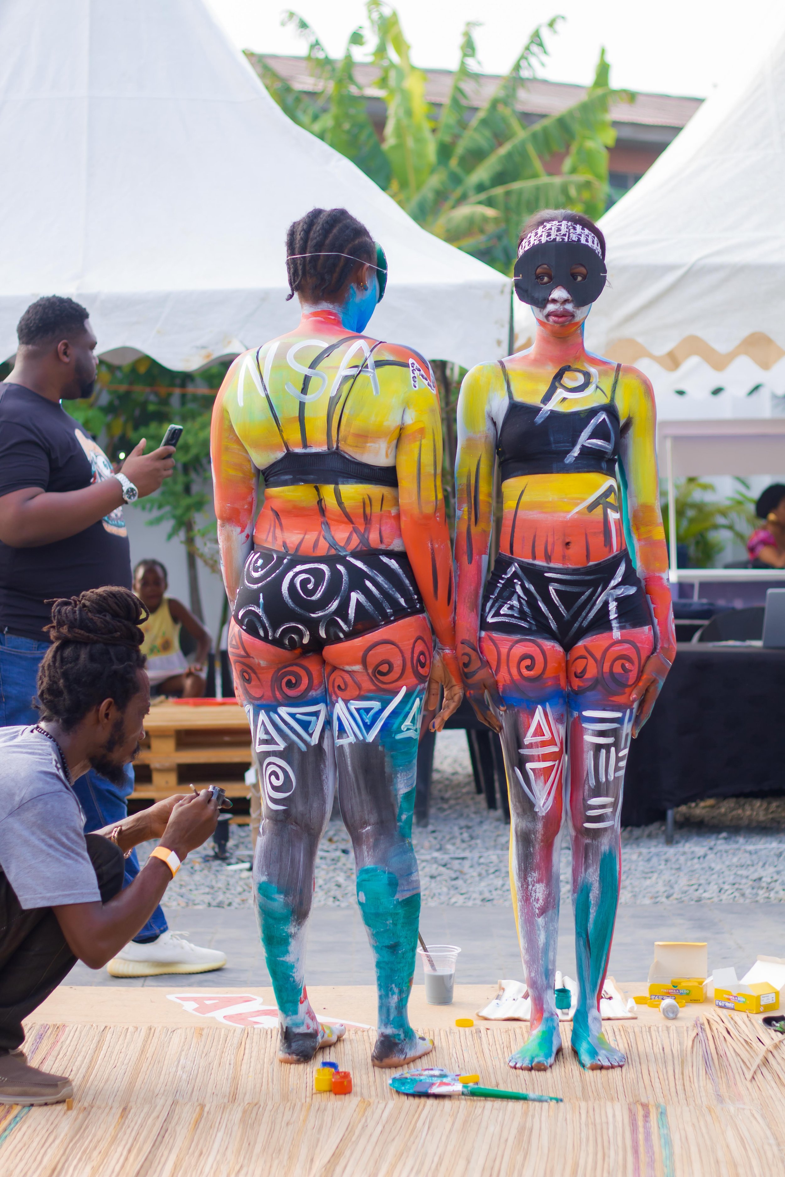 Two women with bodies painted in colorful abstract patterns, standing back-to-back, with the woman on the right wearing a mask, at an outdoor event.