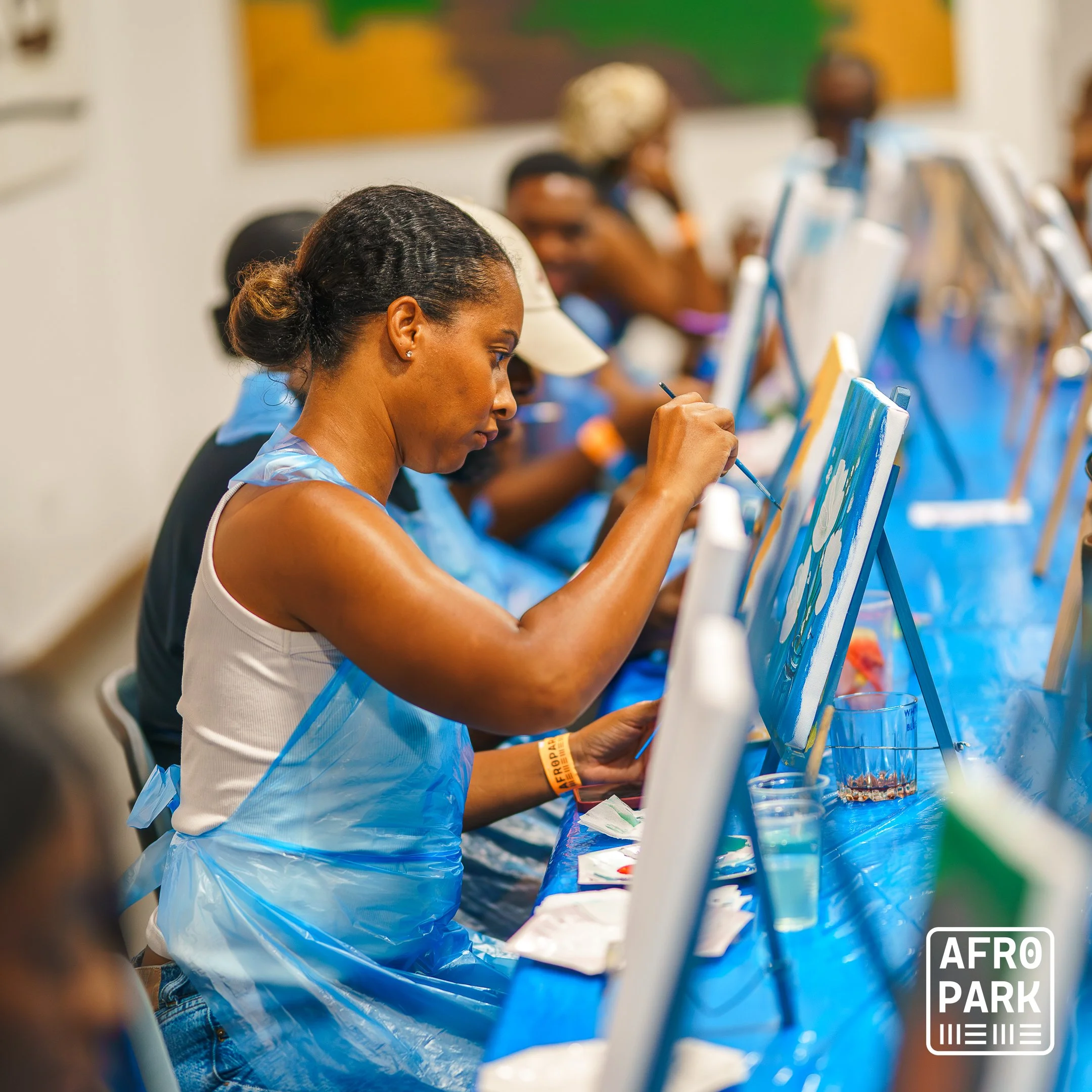 A woman wearing a white tank top, a wristband, and a plastic apron painting on a canvas at an art workshop. Other participants are visible in the background, all seated at a long table with art supplies.
