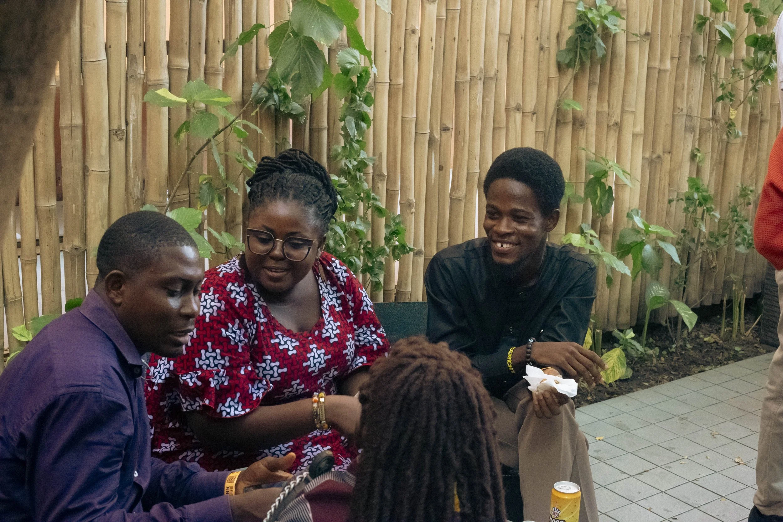 A group of four young people sitting together outside in front of a bamboo fence, engaged in conversation and smiling, with a woman in the foreground and a man in the background holding a tissue.