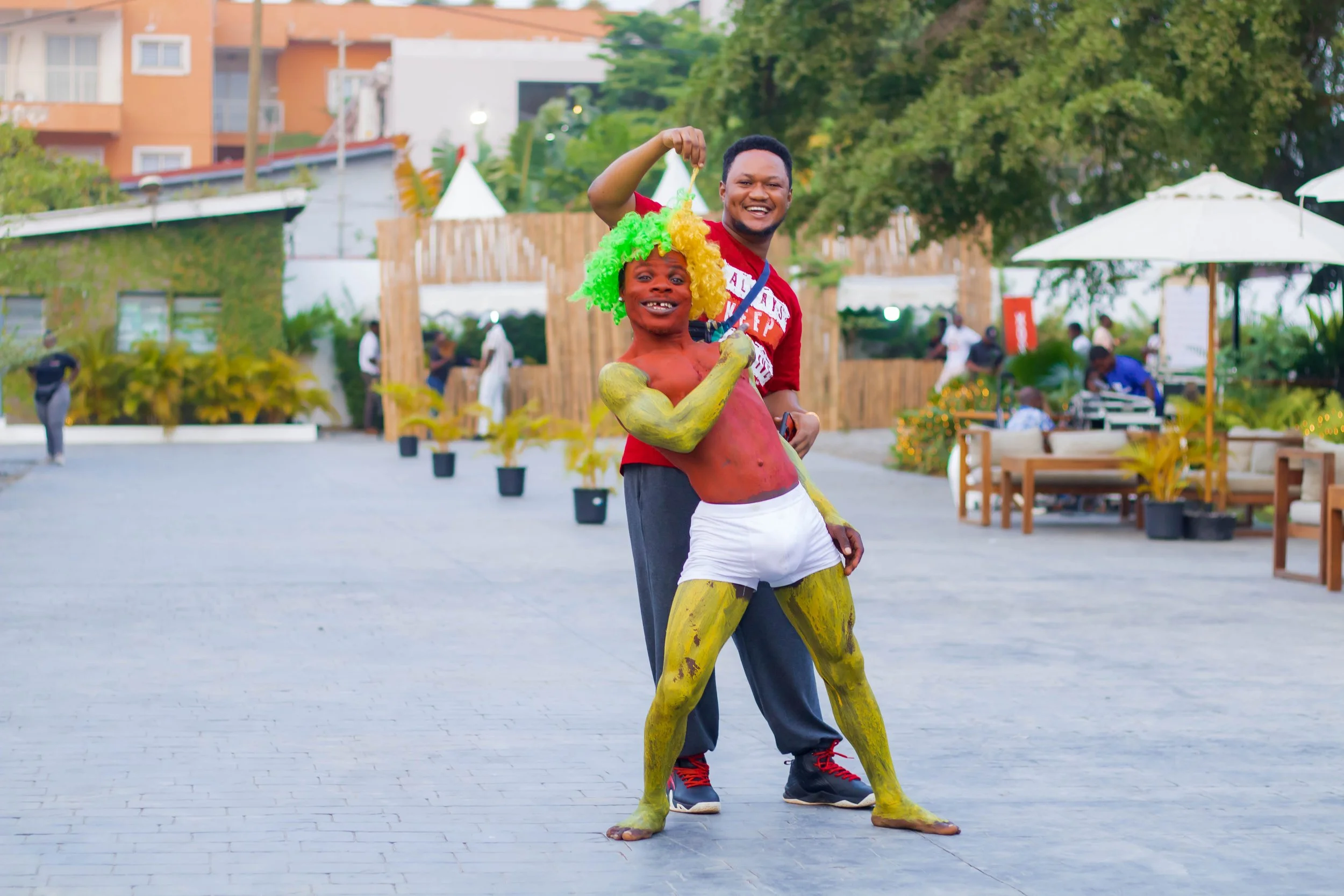 A man standing behind a person dressed in a painted body costume with green hair, yellow painted arms and legs, red chest, white shorts, posing with a smiling man holding a small object, in an outdoor setting with trees, umbrellas, and people in the 
