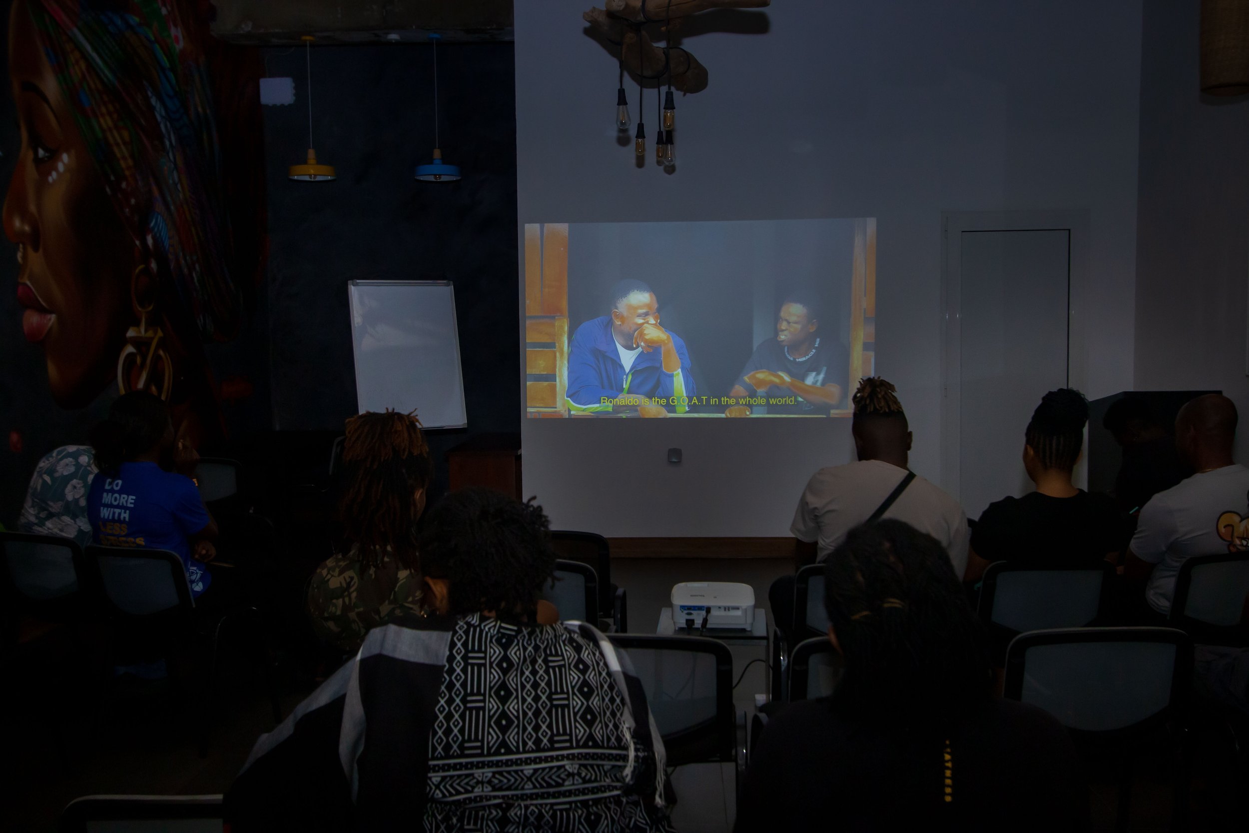 People seated in a dark room watching a video presentation projected onto a wall, featuring two men and yellow subtitles.