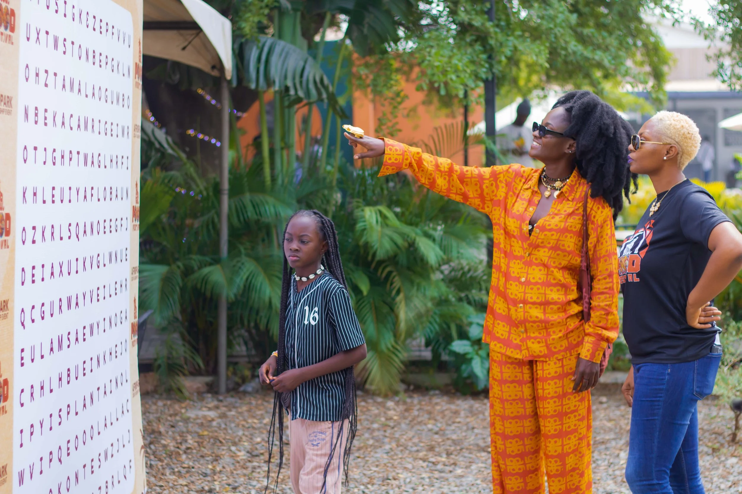 Three women and one young girl standing outdoors near a large eye chart with alphabet letters. The woman in the middle, wearing orange patterned outfit, is pointing at the chart. The young girl, wearing a striped shirt and pink pants, looks on with a
