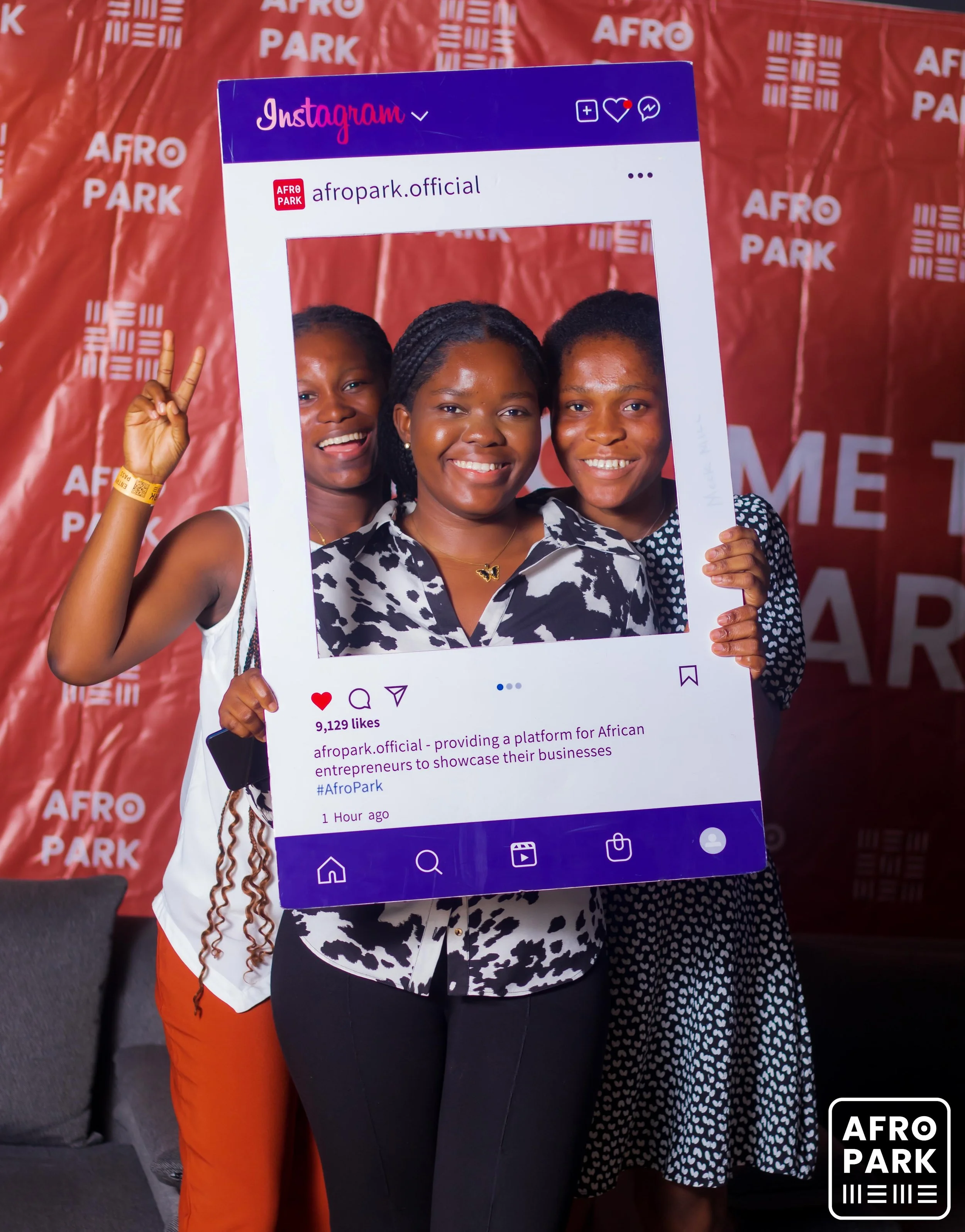 Three women holding a large Instagram frame at an Afro Park event, with a red backdrop displaying 'AFRO PARK' logos. They are smiling and posing for the photo.