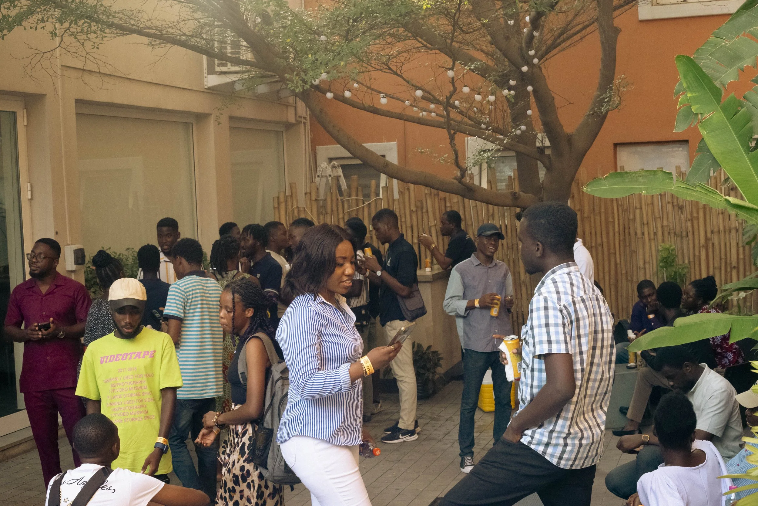 A social gathering of young adults outdoors with some talking, seated, and holding drinks. There is a large tree with string lights, a wooden fence, and greenery in the background.