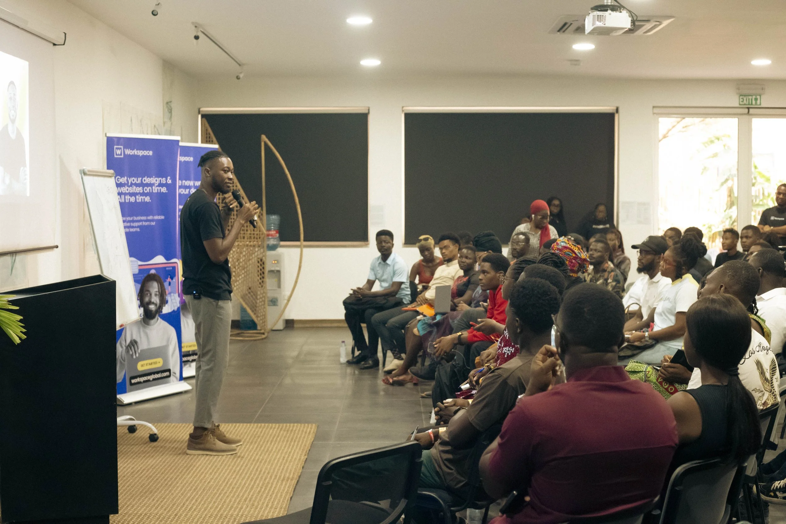 A speaker in a black shirt and beige pants stands on a small stage with a microphone, addressing an audience seated in a conference room. The audience is diverse, listening attentively, some taking notes, with large windows and blackboards behind the