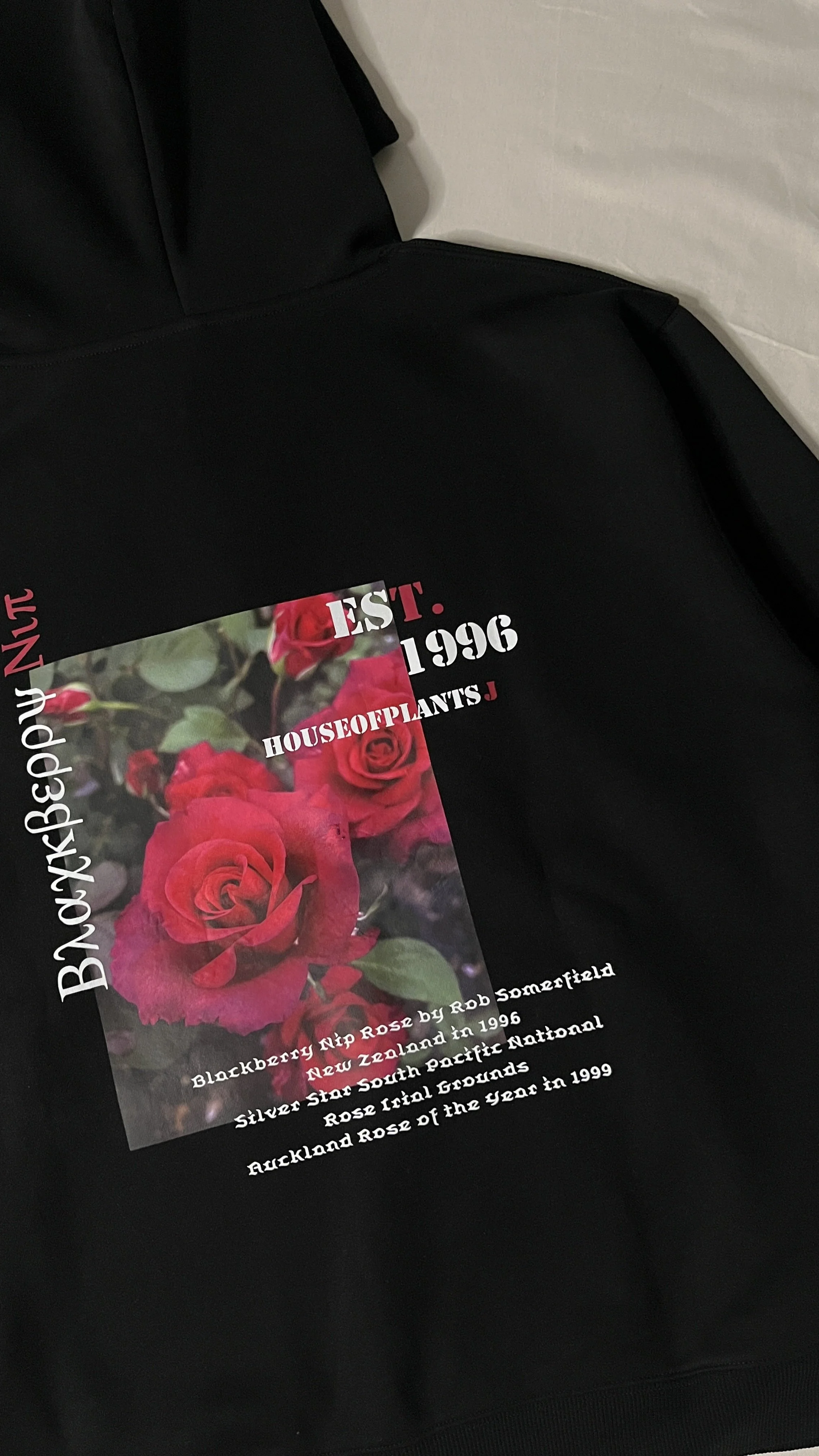 Black hoodie with a printed graphic of red roses and text about houseplants, including information about Rob Somerfield and the Auckland Rose of the Year in 1999.