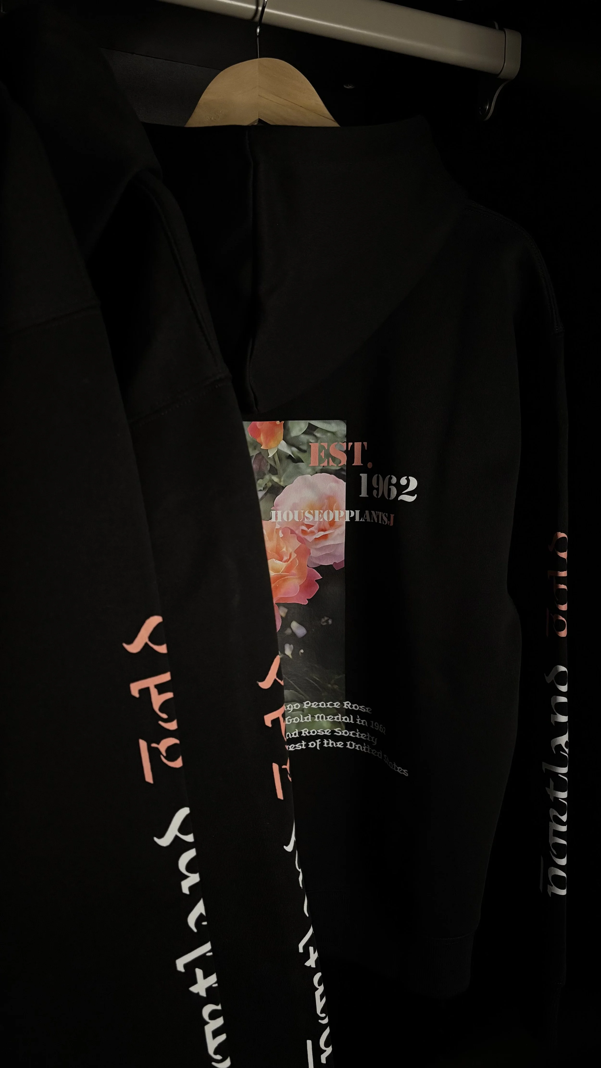 Black hoodie hanging on a wooden hanger. The hoodie features a floral design and text referencing the establishment of a house or organization in 1962.