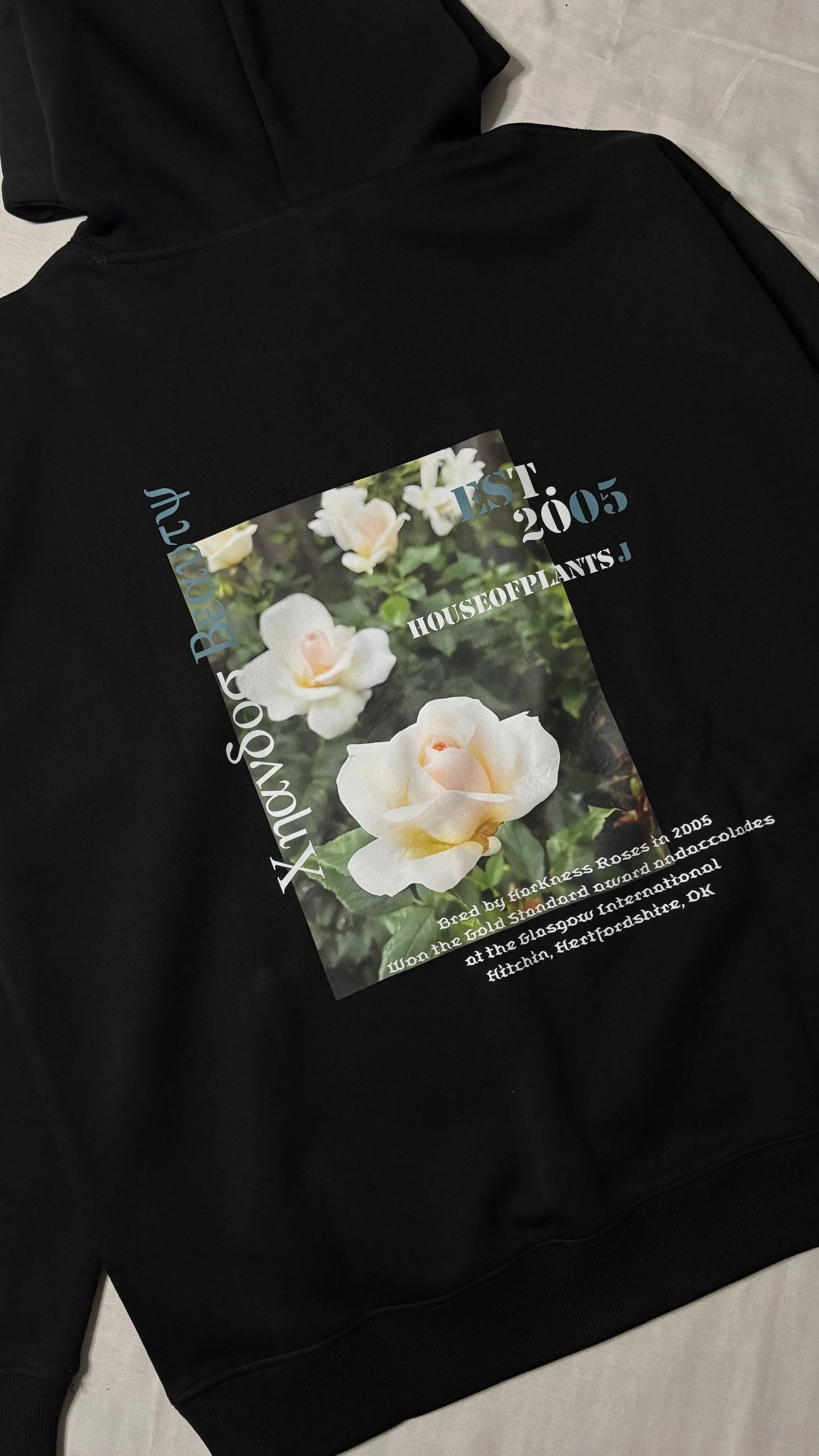 Black hooded sweatshirt with a large printed photo of white roses and text about a 2005 rose breed from Hertfordshire, UK.