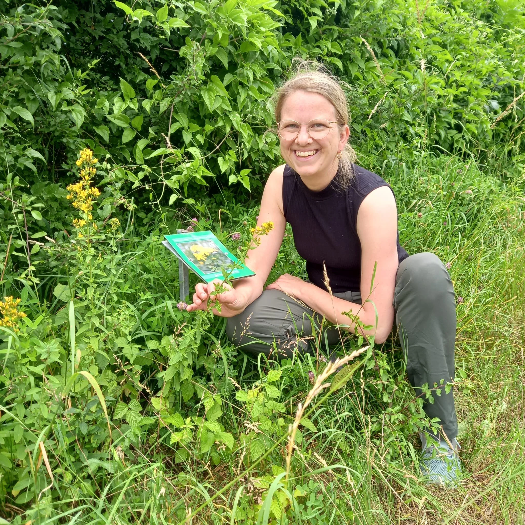 Frau sitzt im Grünen, hält eine Plakette mit Pflanzenbild, umgeben von diversen grünen Pflanzen und Blumen.
